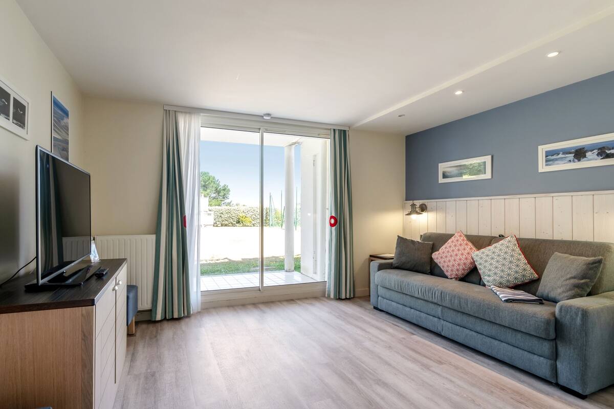 Una sala de estar moderna con un televisor de pantalla plana, un sofá gris con cojines estampados y un ventanal corredizo que da a un área exterior.