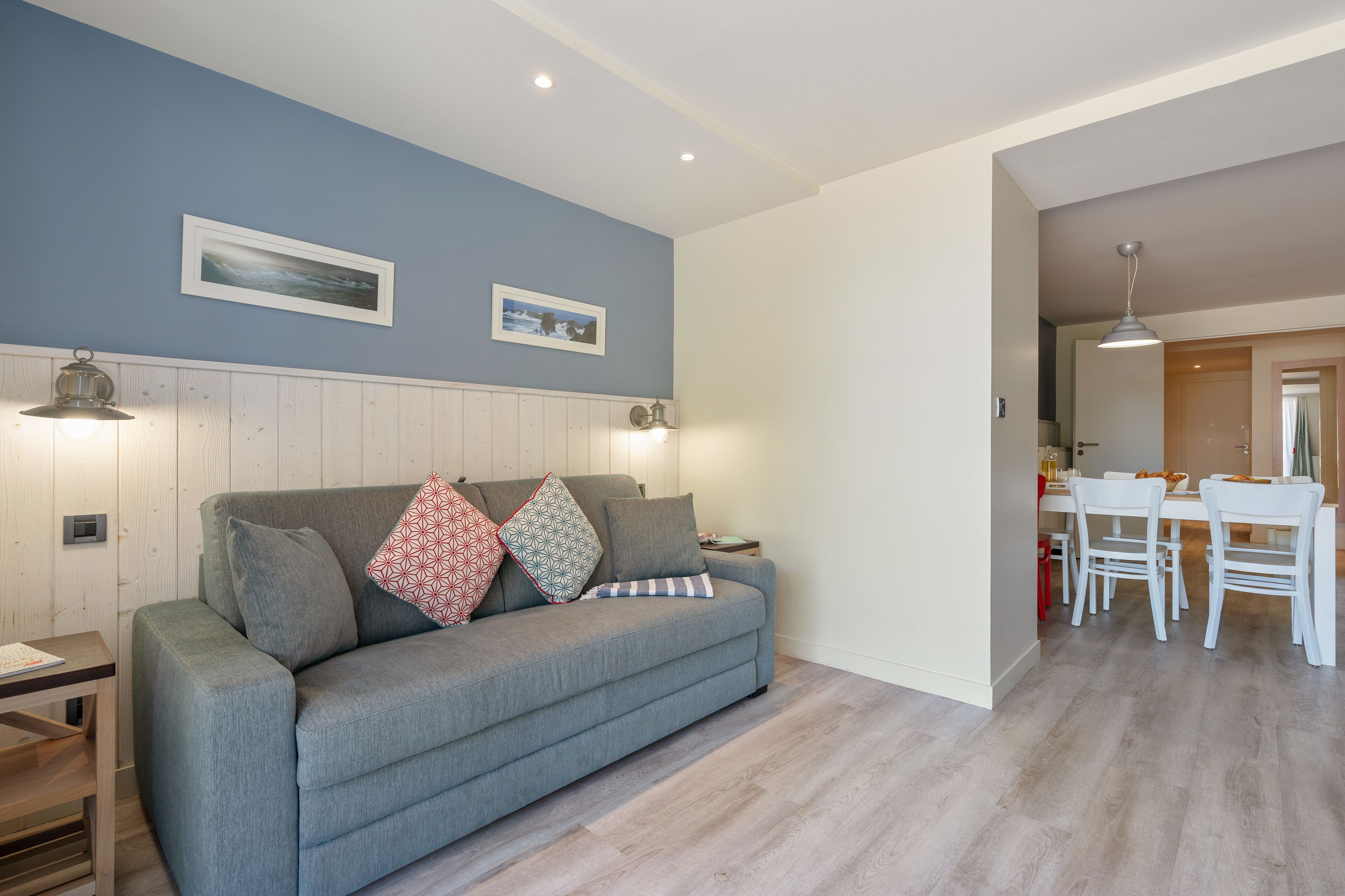 Un salón con un sofá gris, dos lámparas de pared y una zona de comedor con mesa y sillas.