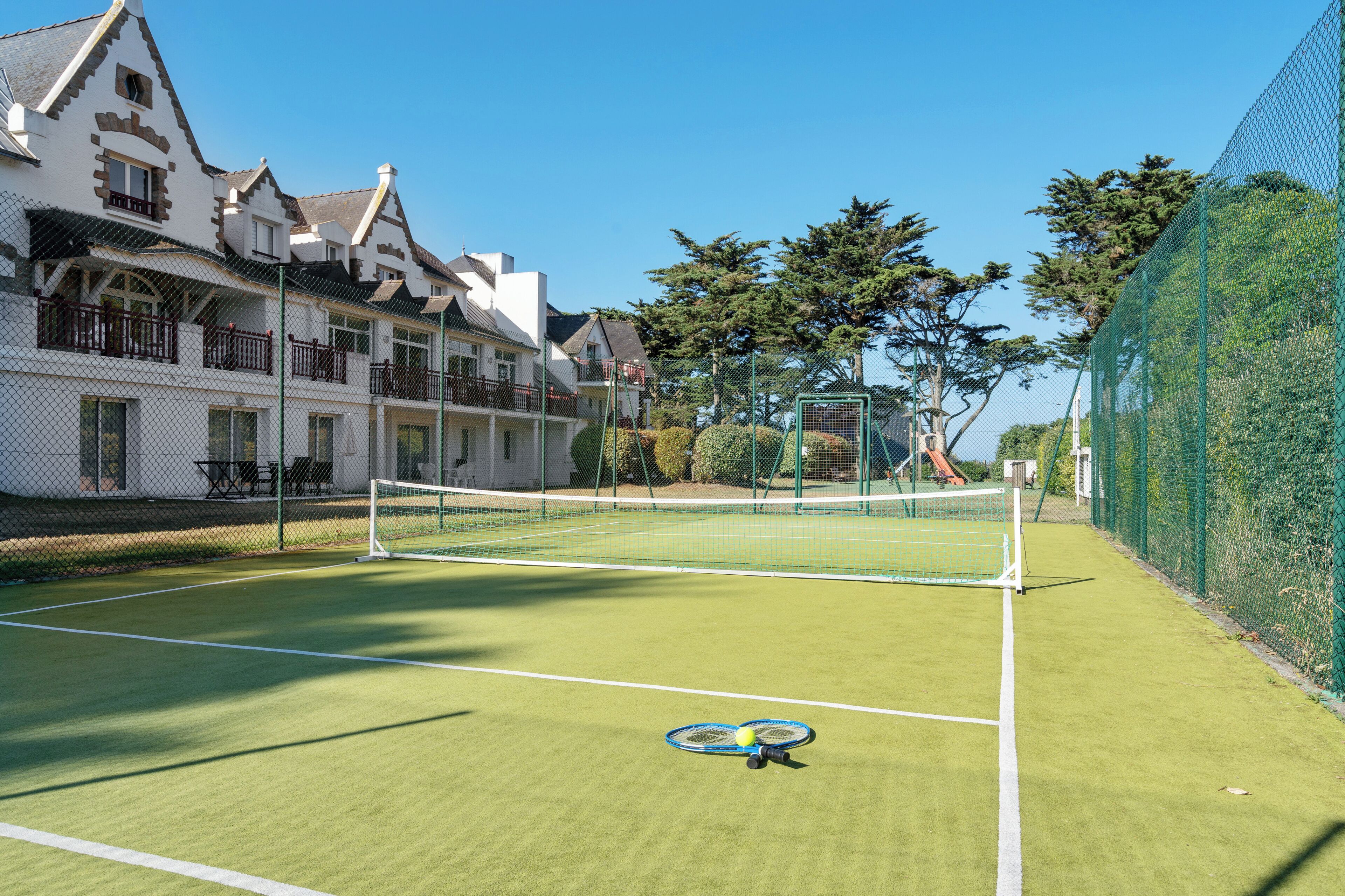 Een tennisbaan met een net, omringd door een groene hek en een wit gebouw met meerdere balkons.