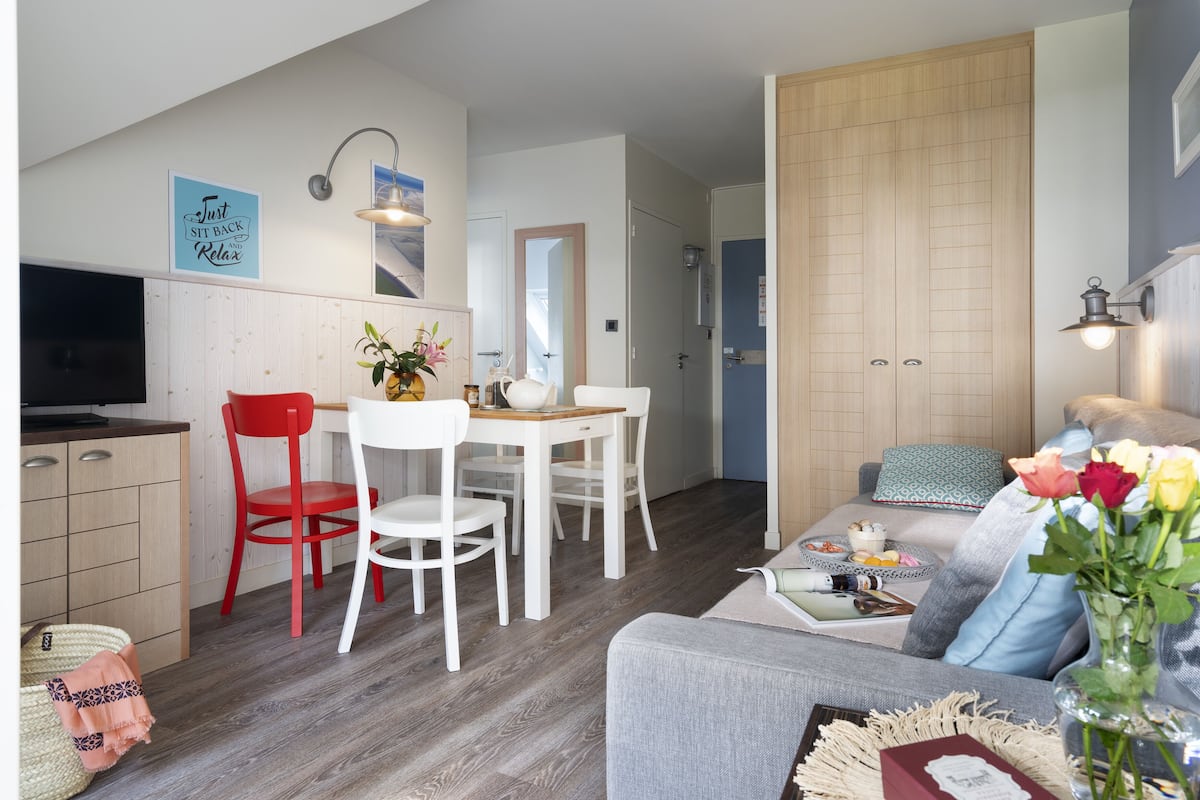 Una sala moderna con un sofá gris, una silla roja, una mesa de comedor con sillas blancas y una zona de cocina con armarios de madera y un televisor.