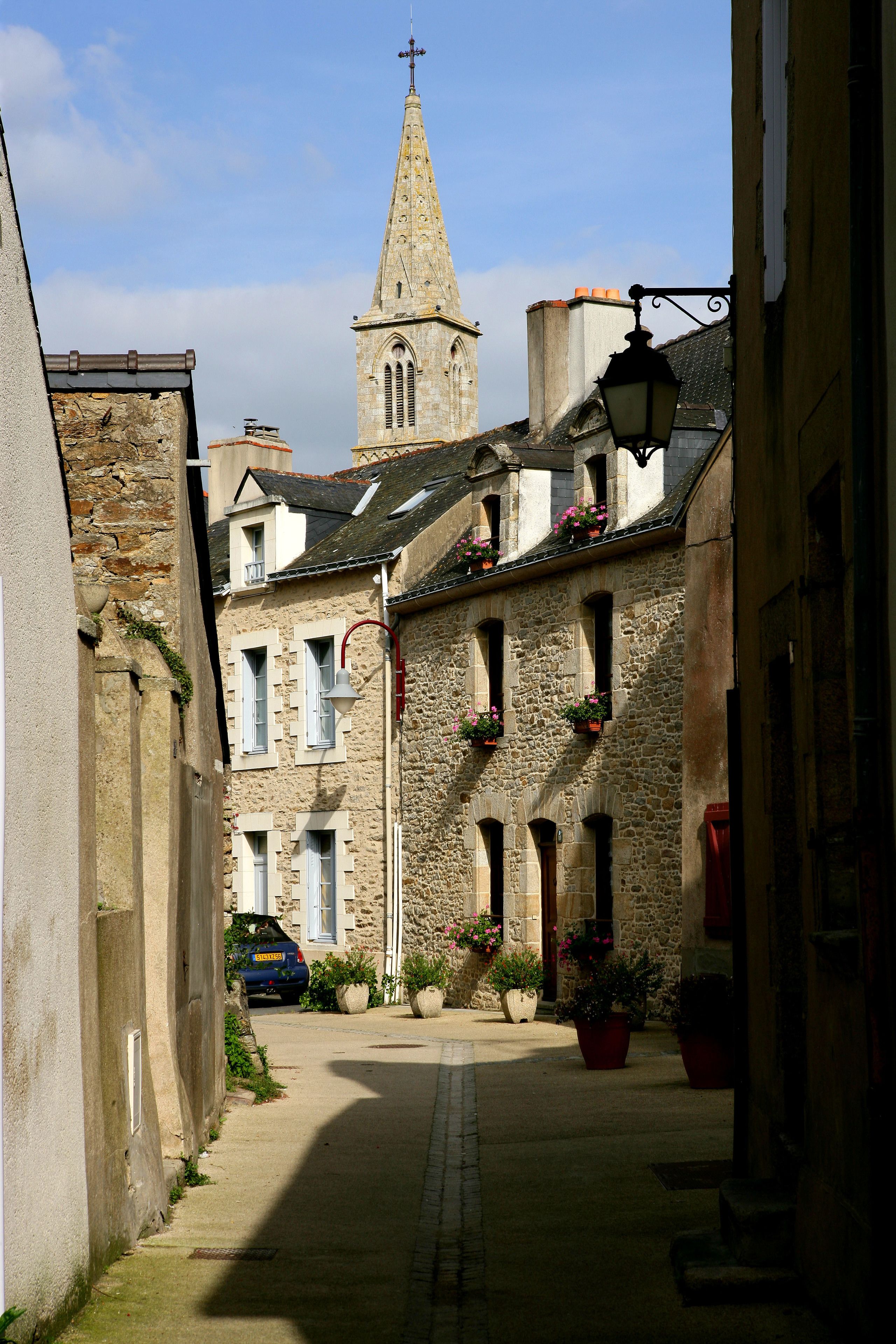 A narrow street with stone buildings, a church tower, and a parked car.