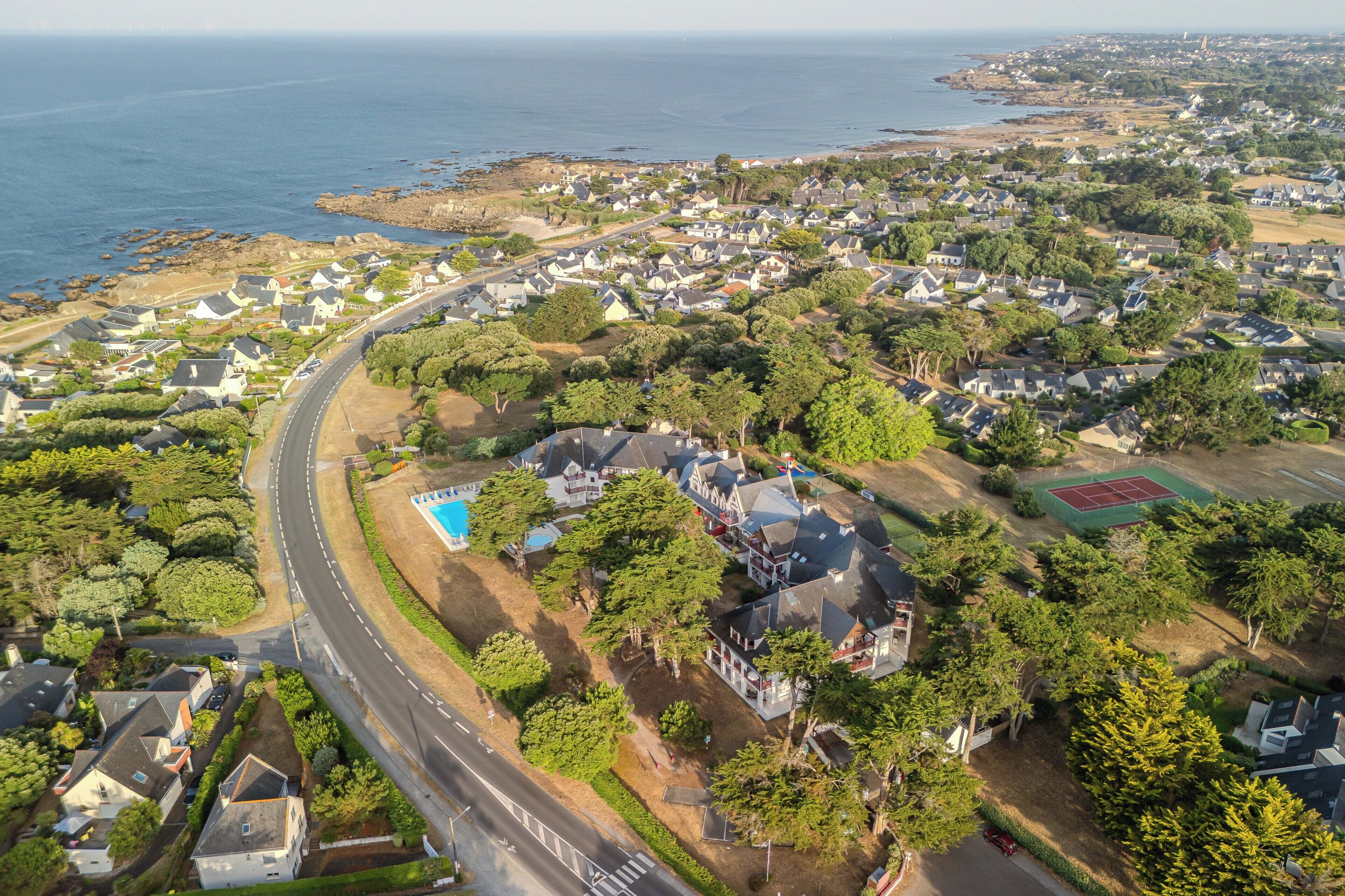 Vue aérienne d’un quartier résidentiel situé sur la côte, avec des maisons, une route et un court de tennis.