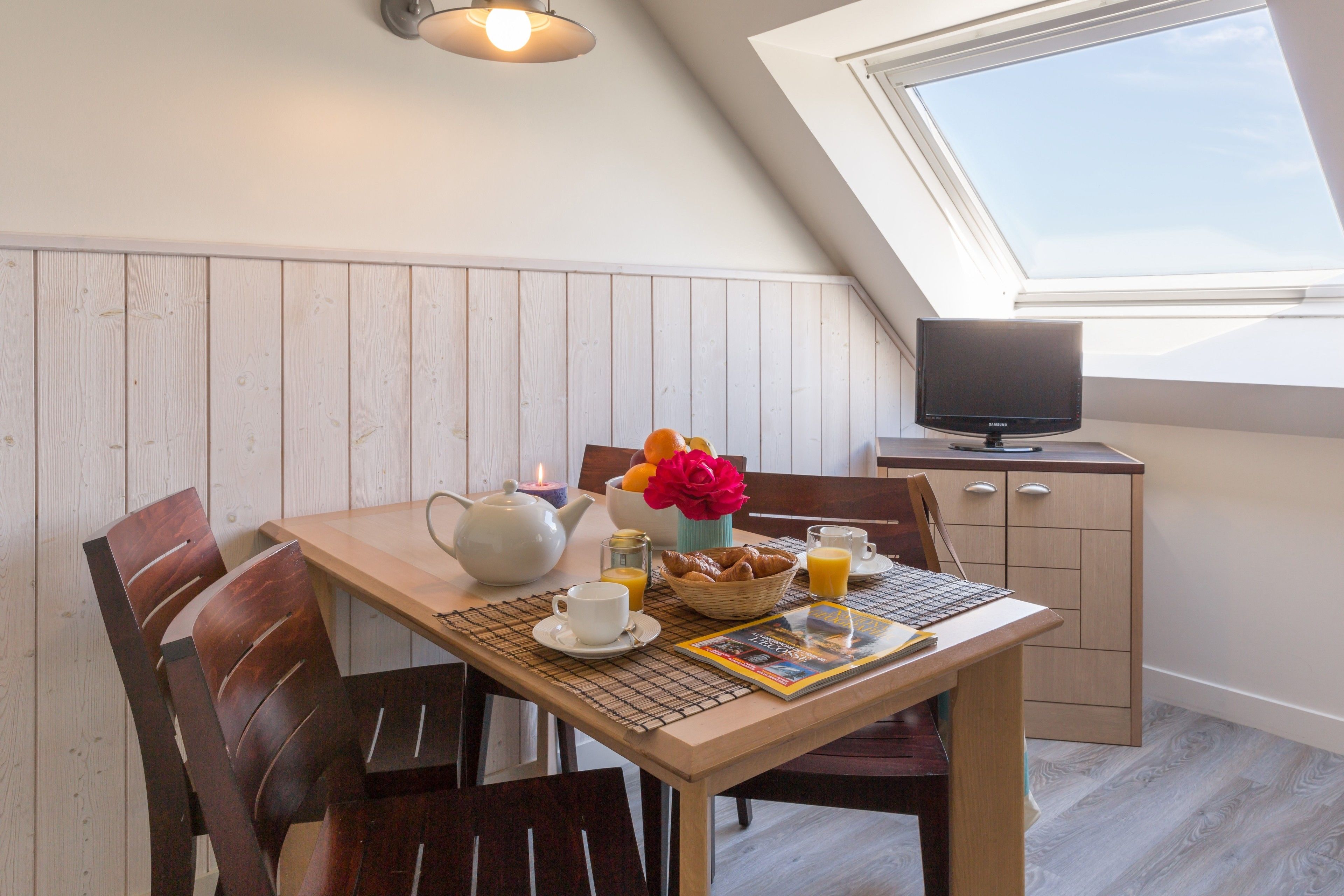 Un área de comedor con una mesa dispuesta para el desayuno, un televisor sobre un mueble bajo y un tragaluz arriba.