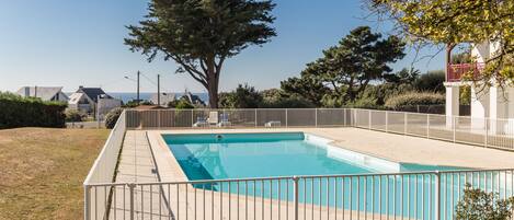 A rectangular swimming pool with a white fence, surrounded by grass and trees, with houses in the background.