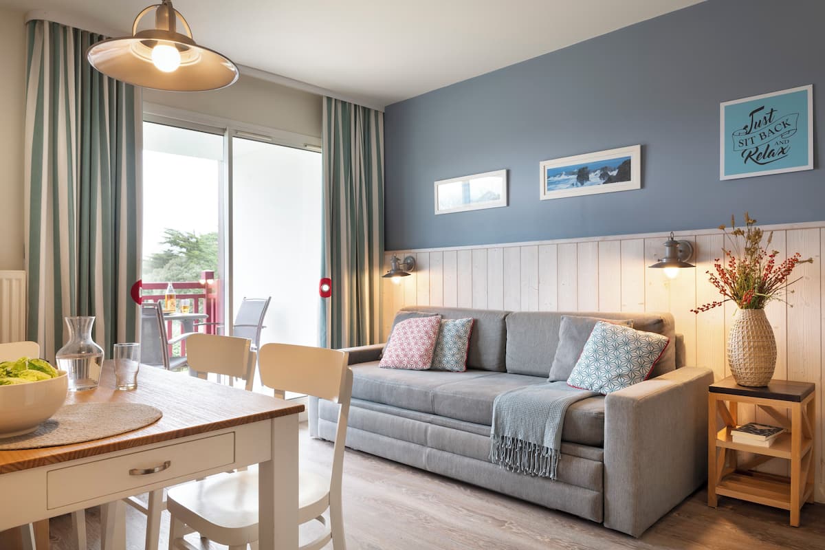 Una sala de estar acogedora con un sofá gris, una mesa de centro de madera y una mesa de comedor con sillas. Hay un ventanal con cortinas, una lámpara de pared y un cuadro enmarcado.