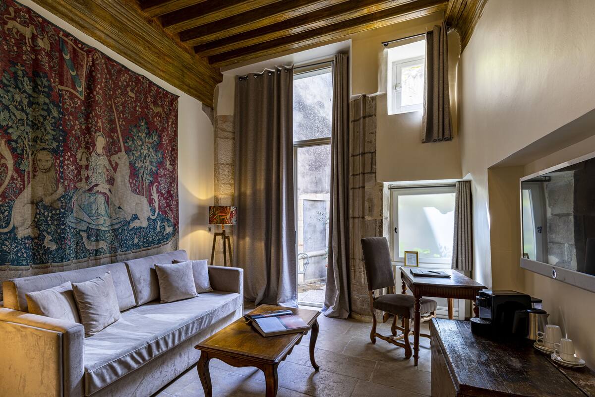 Een gezellige woonkamer met een groot wandtapijt, een bank, een houten salontafel, een stoel en een klein tafeltje met een koffieapparaat.