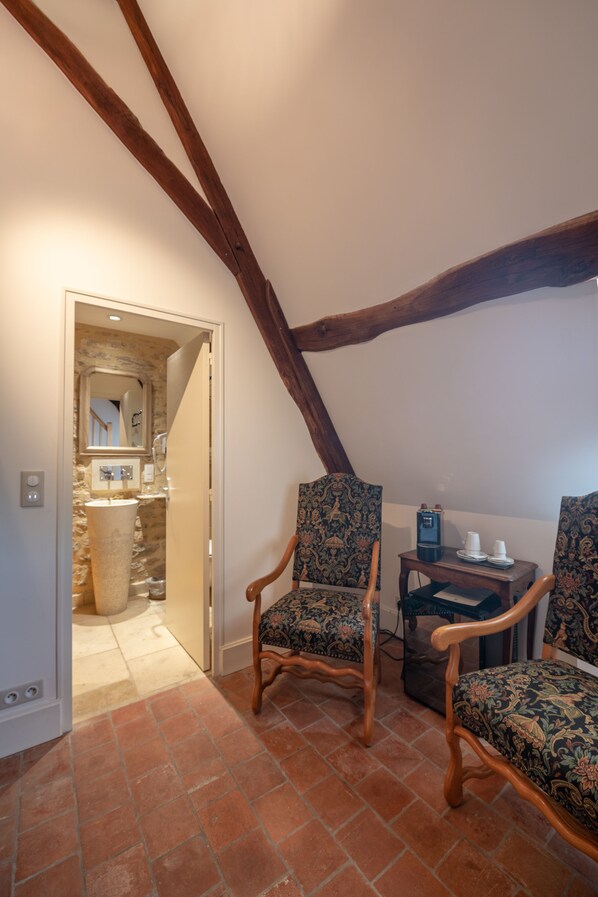 A room with a sloped ceiling, wooden beams, two patterned chairs, a small table with cups and a coffee maker, and a doorway leading to a bathroom with a stone sink.