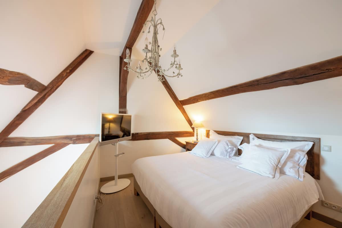 Een slaapkamer met een houten plafond, een bed met wit beddengoed, een nachtkastje met een lamp en een vloerlamp.