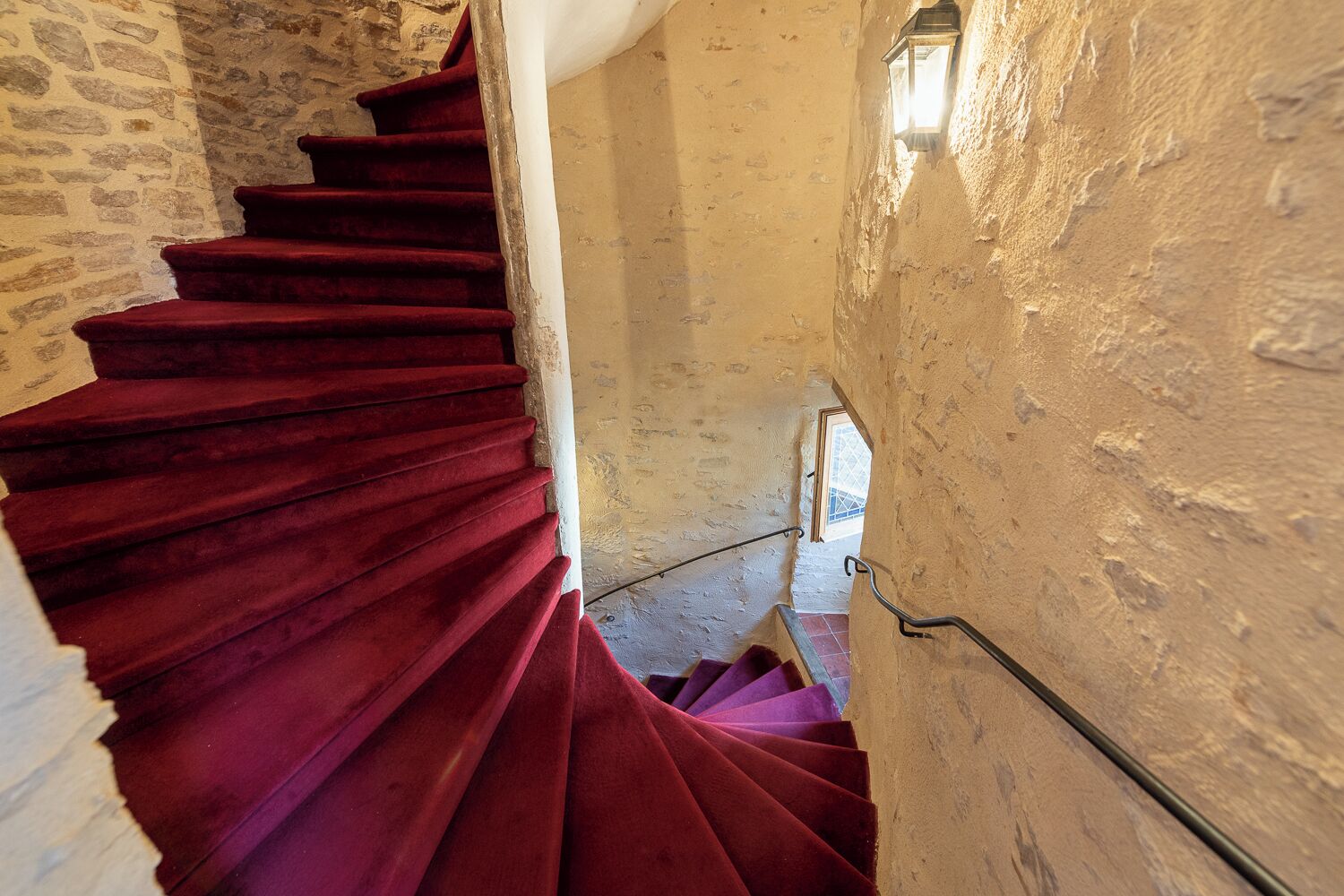 Un escalier en colimaçon, dont les marches sont recouvertes d’une moquette rouge, mène à un niveau inférieur.