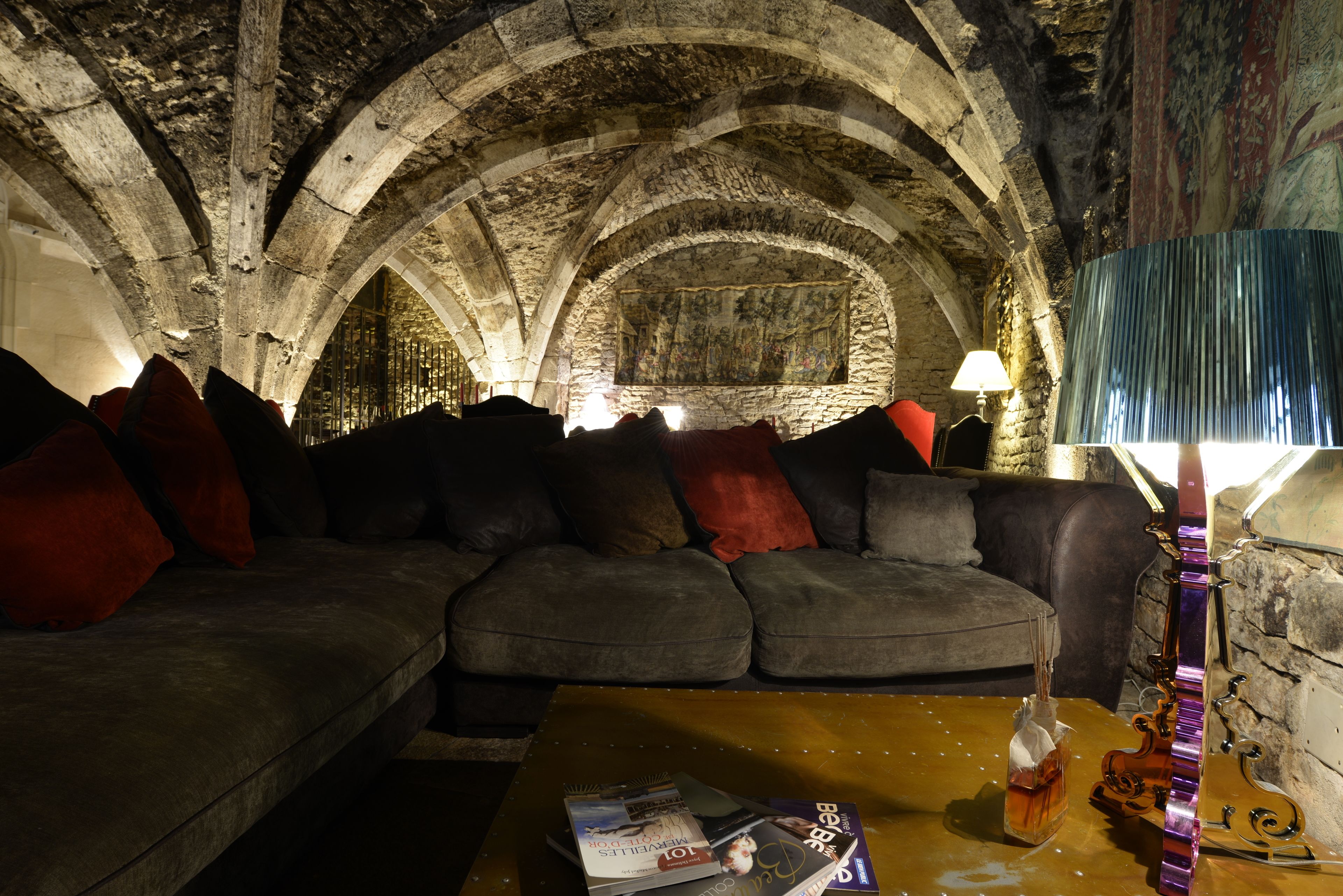 Un salon chaleureux avec un plafond en pierre, un canapé sombre agrémenté de coussins rouges, une table basse avec des livres et une bouteille, et un violon appuyé contre le mur.