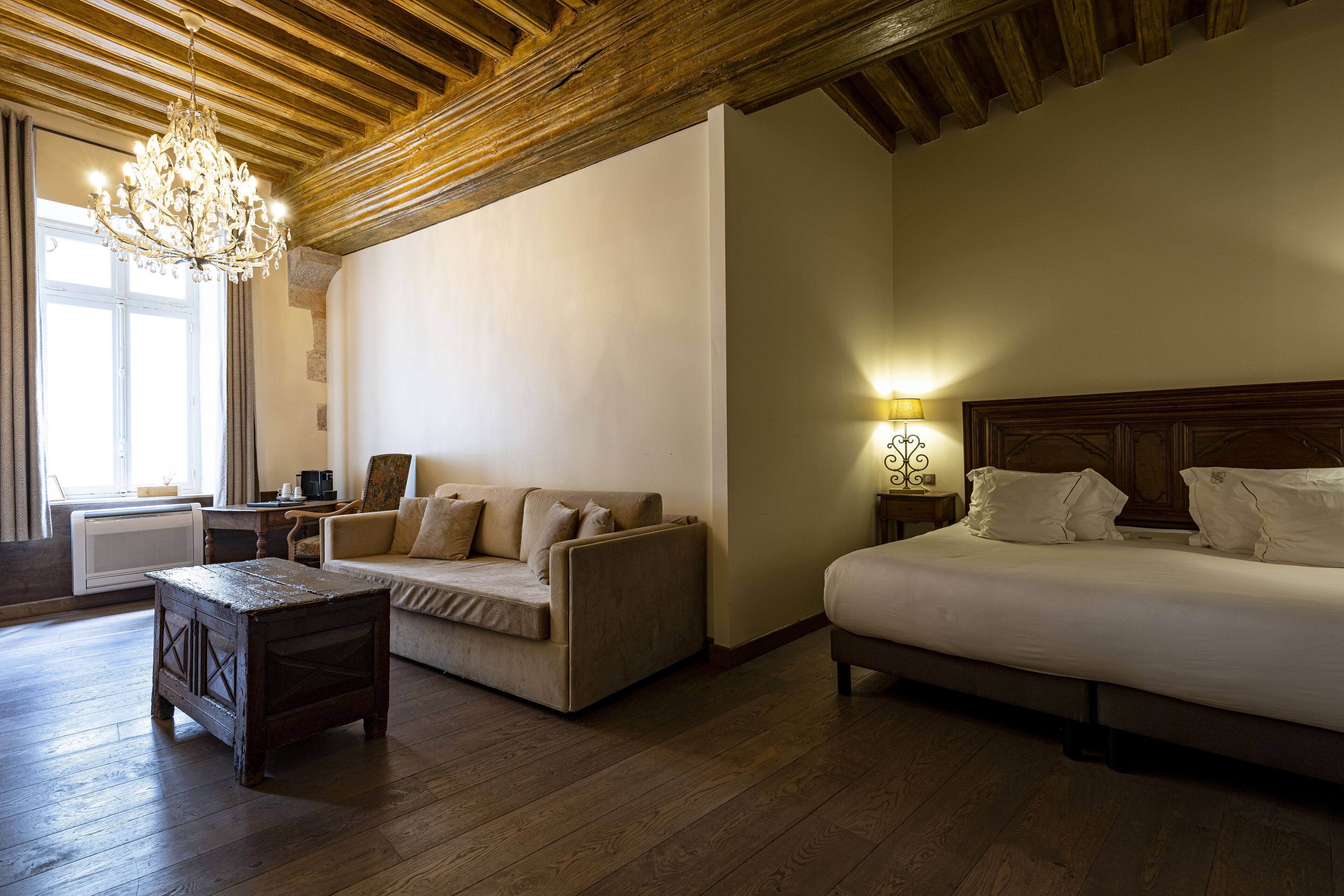 Une chambre d’hôtel avec un plafond en bois, un lustre, un lit, un canapé, une petite table et une table de chevet avec une lampe.