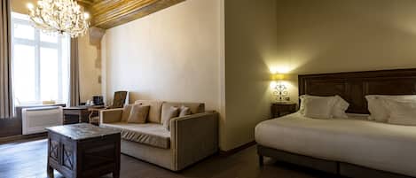 Habitación de hotel con techo de madera, candelabro, cama, sofá, pequeña mesa y mesita de noche con lámpara.