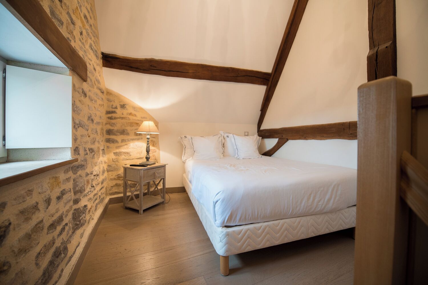 A bedroom with a stone wall, wooden beams, a bed with white bedding, and a nightstand with a lamp.