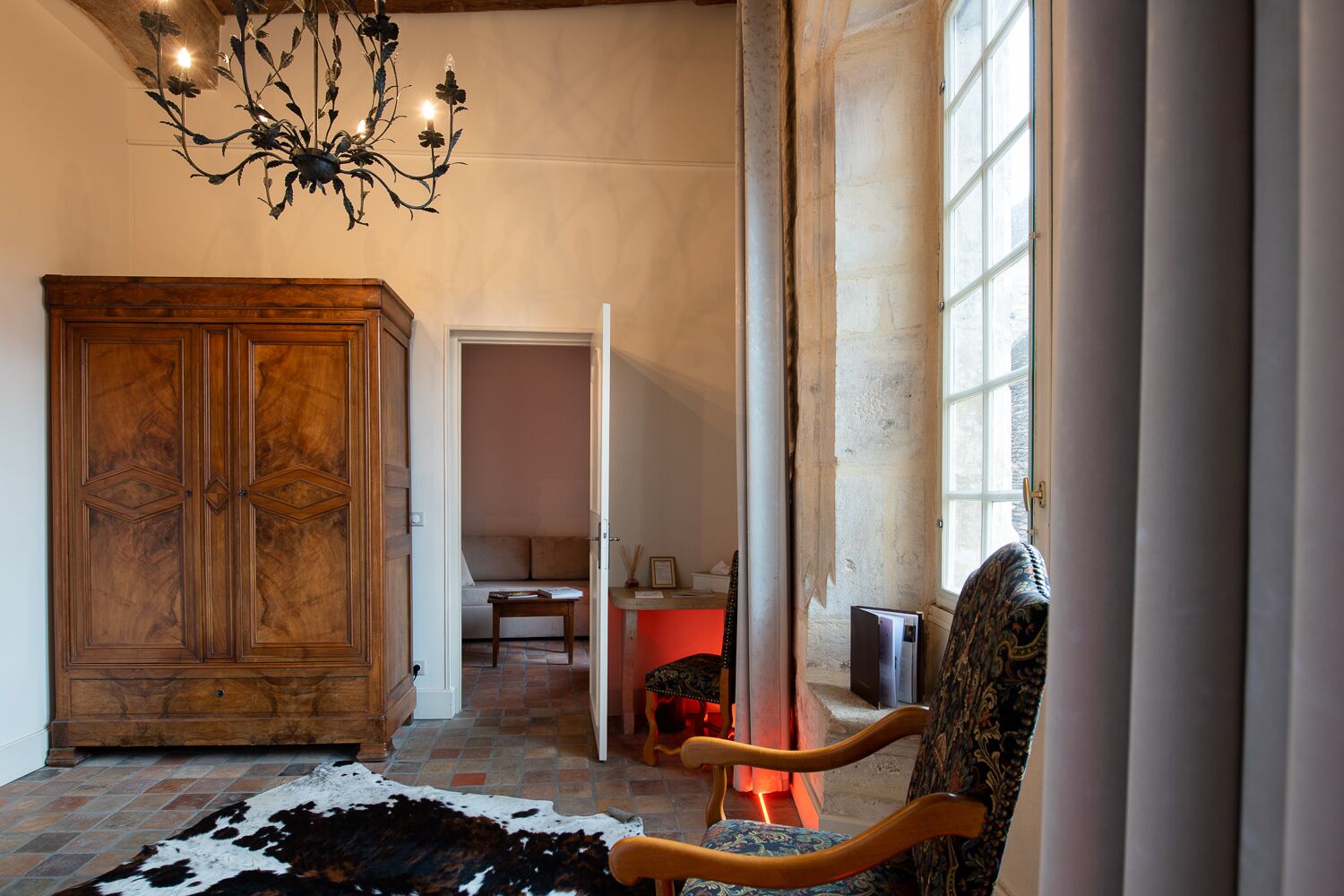 A spacious room with a large wooden cabinet, a patterned armchair, a cowhide rug, and a chandelier.