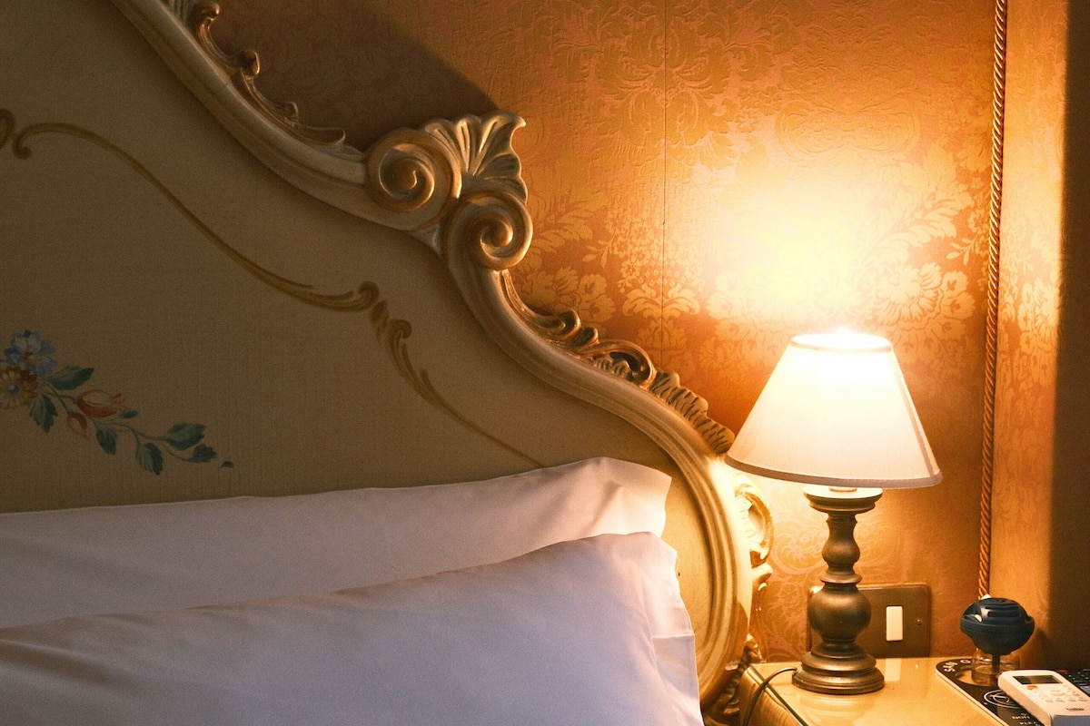 A neatly made bed with white linens, a classic ornate headboard, and a bedside table with a lamp.