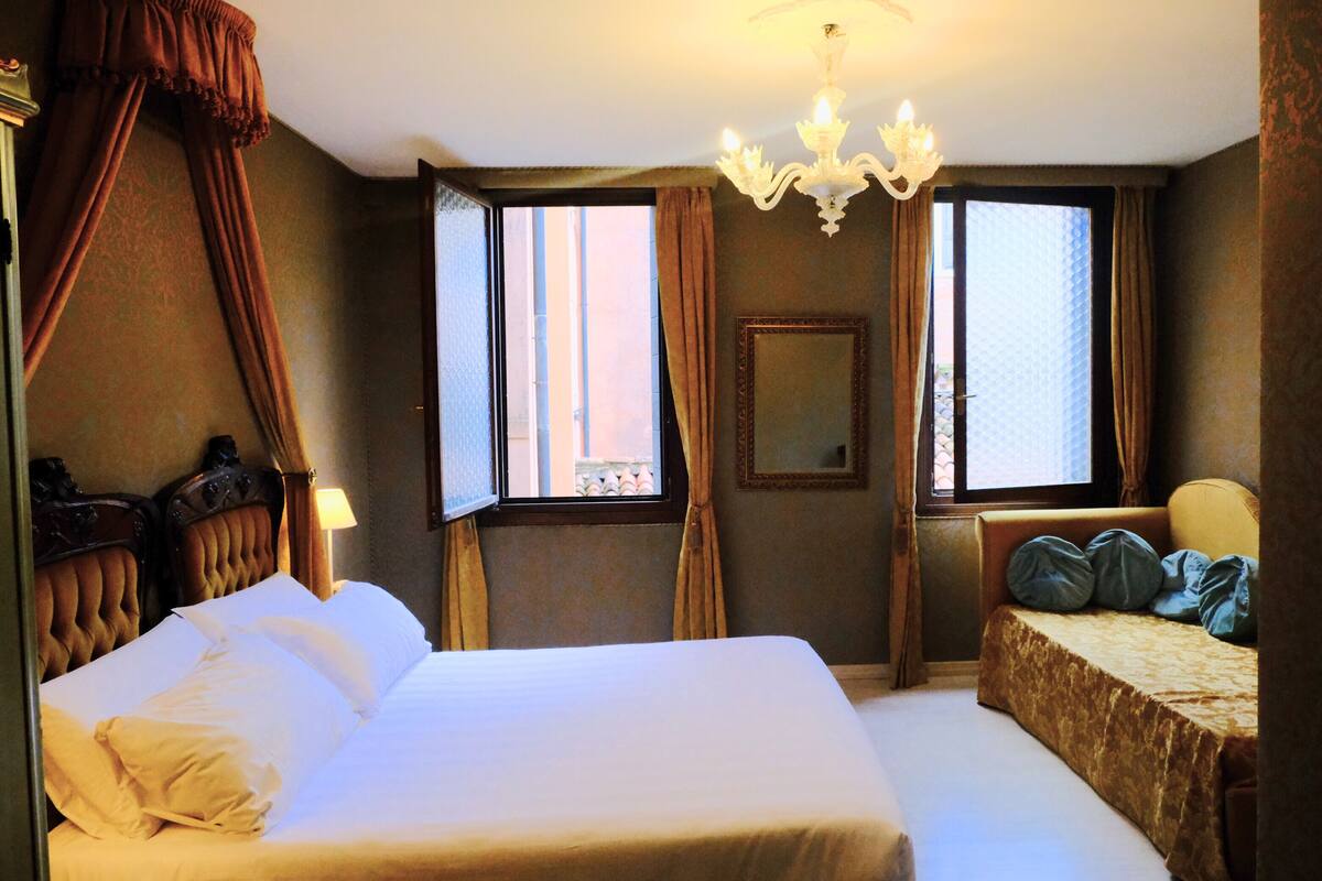 A hotel room with a large bed, a smaller bed, a chandelier, and a window with curtains.