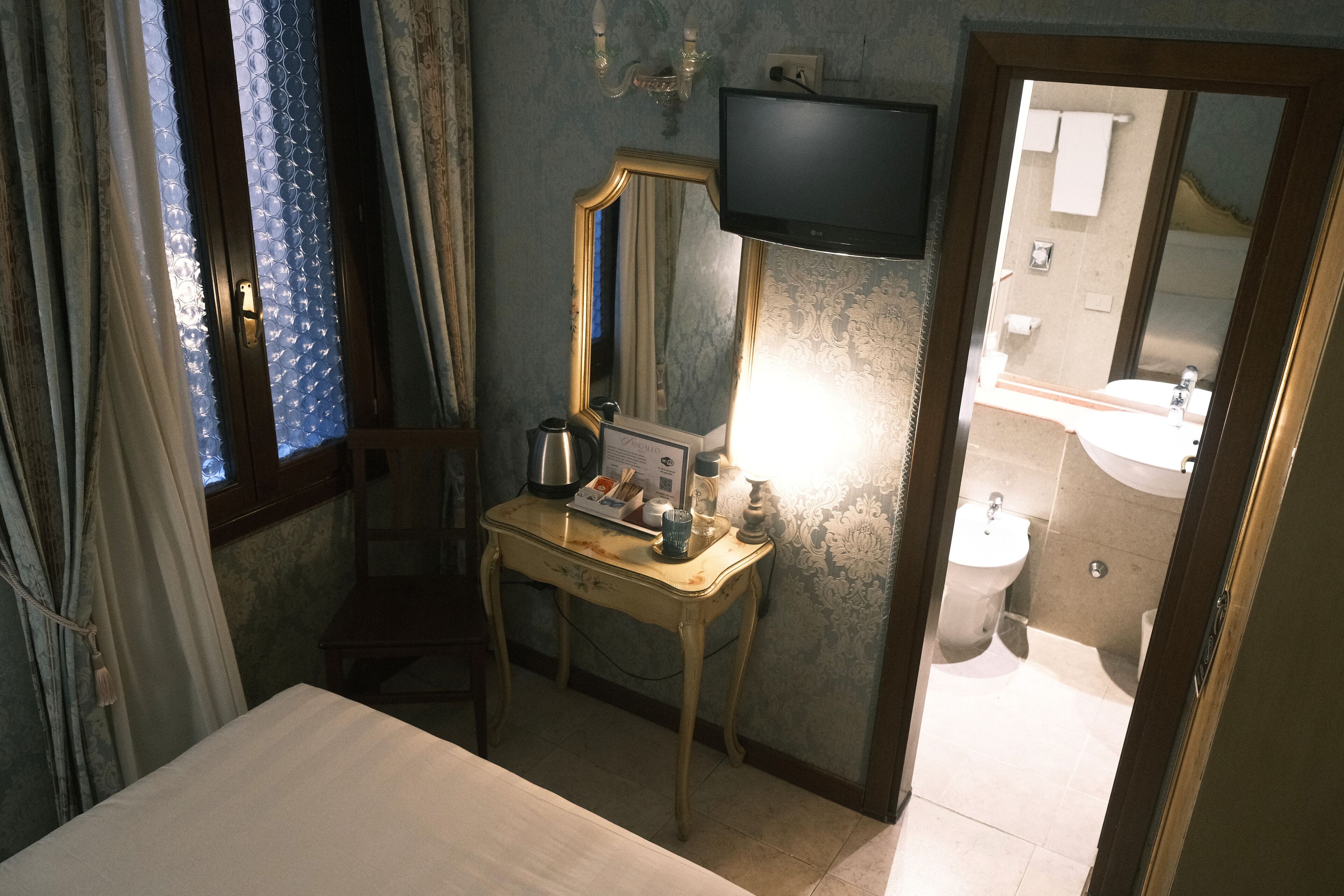 A hotel room with a bed, a desk with a lamp, a television, and a bathroom visible through an open door.