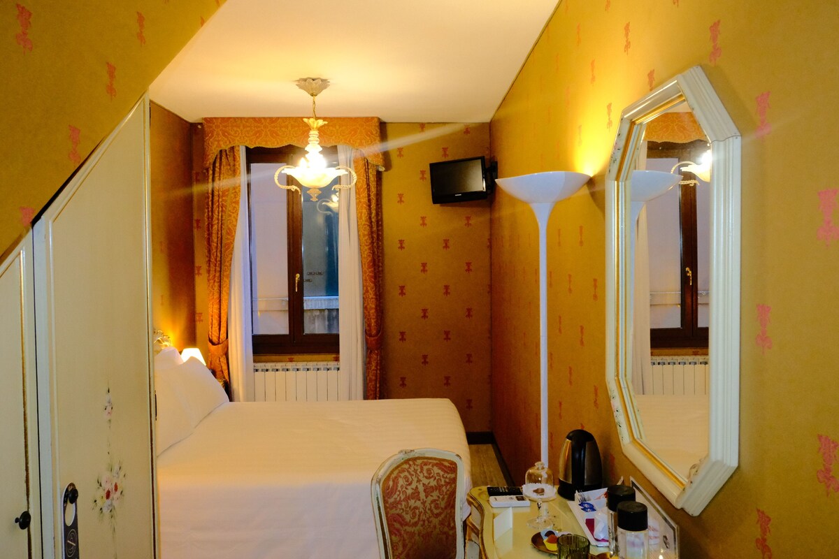 Habitación de hotel con cama, escritorio con lámpara, televisor y espejo grande.