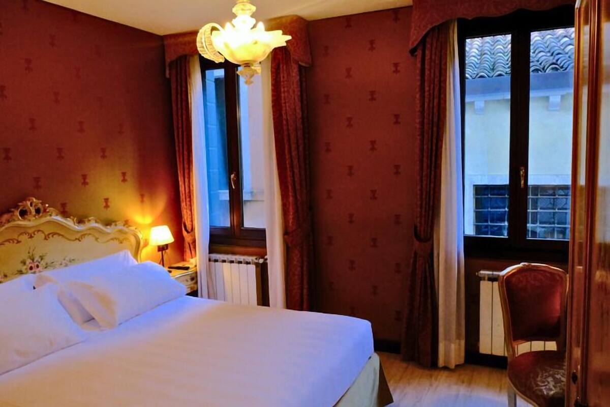 A hotel room with a bed, a chair, a window with curtains, and a chandelier.