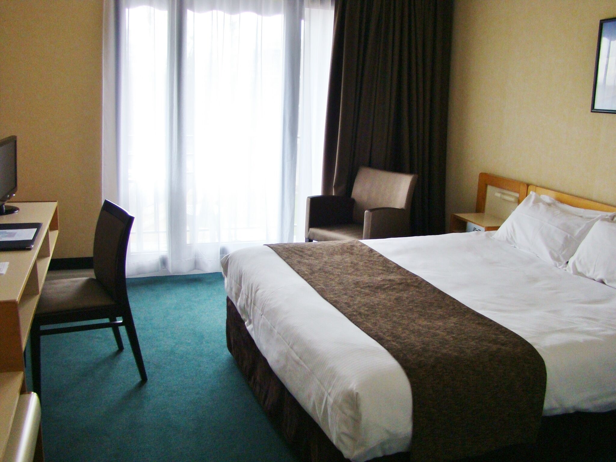 Une chambre d’hôtel comprenant un lit, un bureau avec un ordinateur, une chaise, une table de chevet et une fenêtre avec des rideaux.