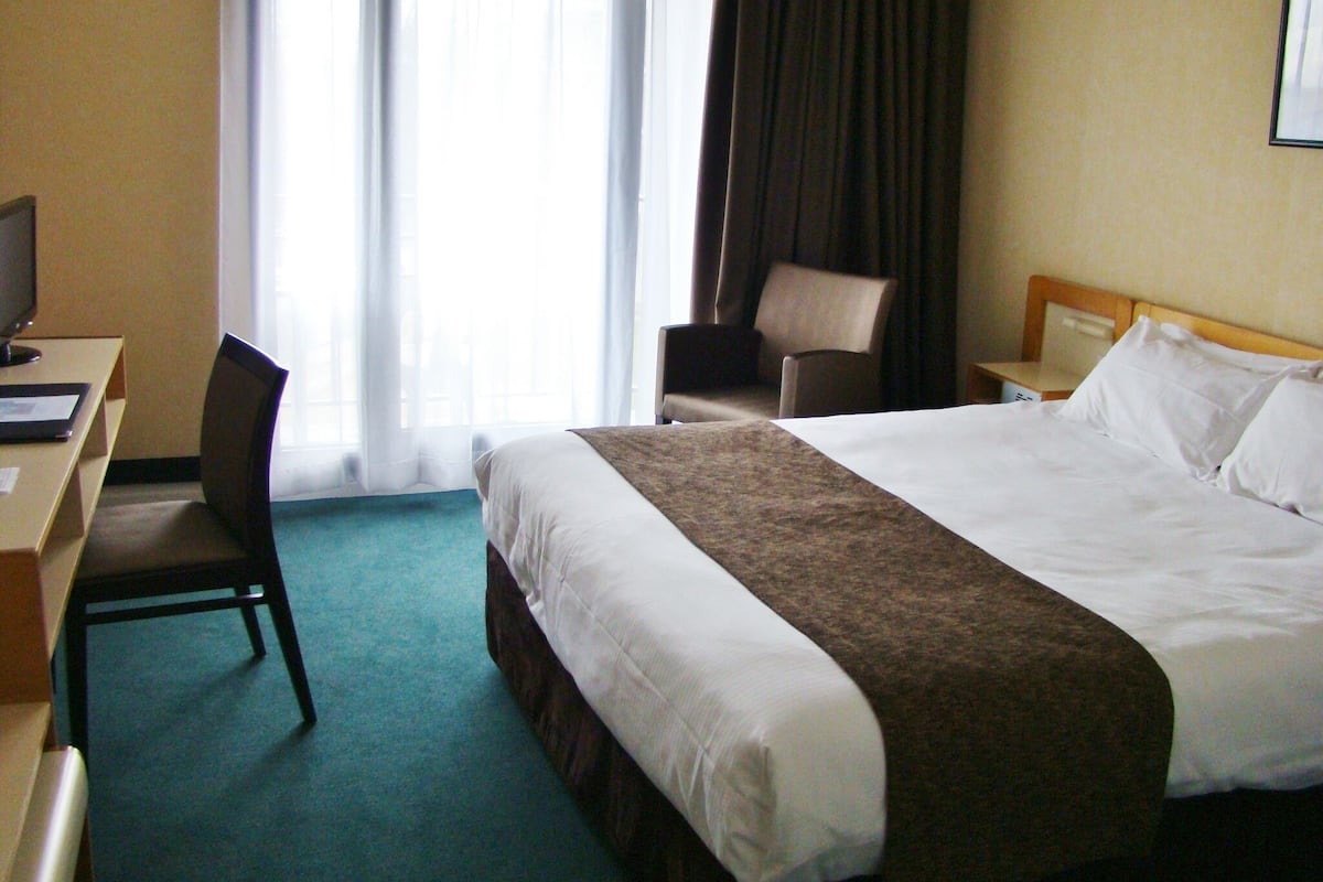 Une chambre d’hôtel comprenant un lit, un bureau avec un ordinateur, une chaise, une table de chevet et une fenêtre avec des rideaux.