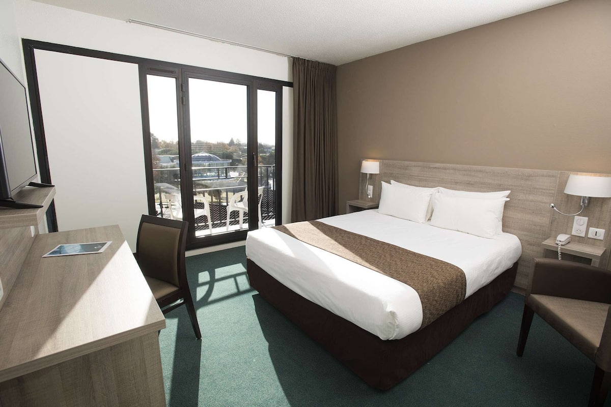 Une chambre d’hôtel avec un grand lit, un bureau et une chaise. La fenêtre offre une vue sur les bâtiments environnants.