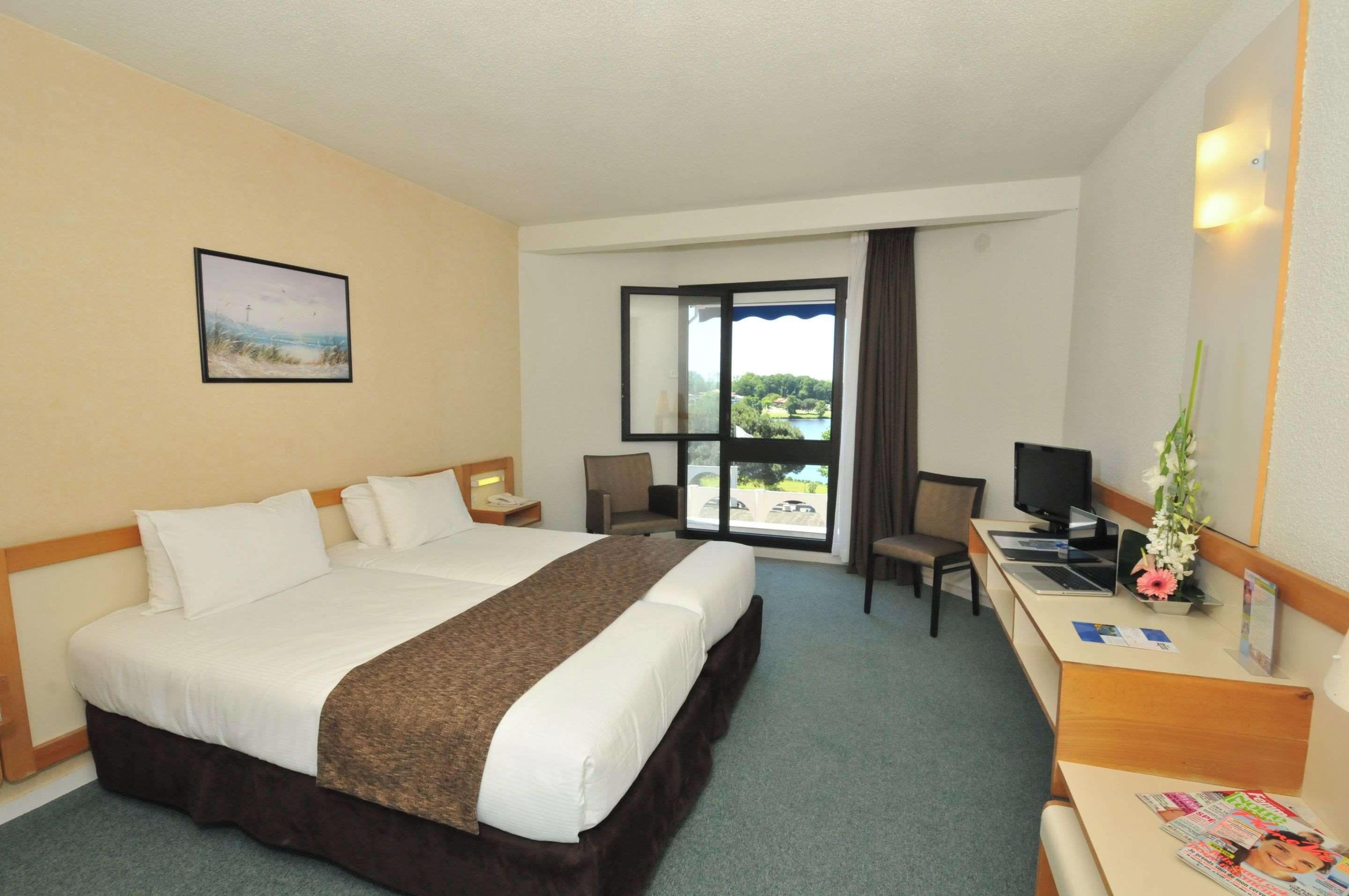 Habitación de hotel con una cama grande, un escritorio con ordenador, una silla y una ventana con vistas a zonas verdes.