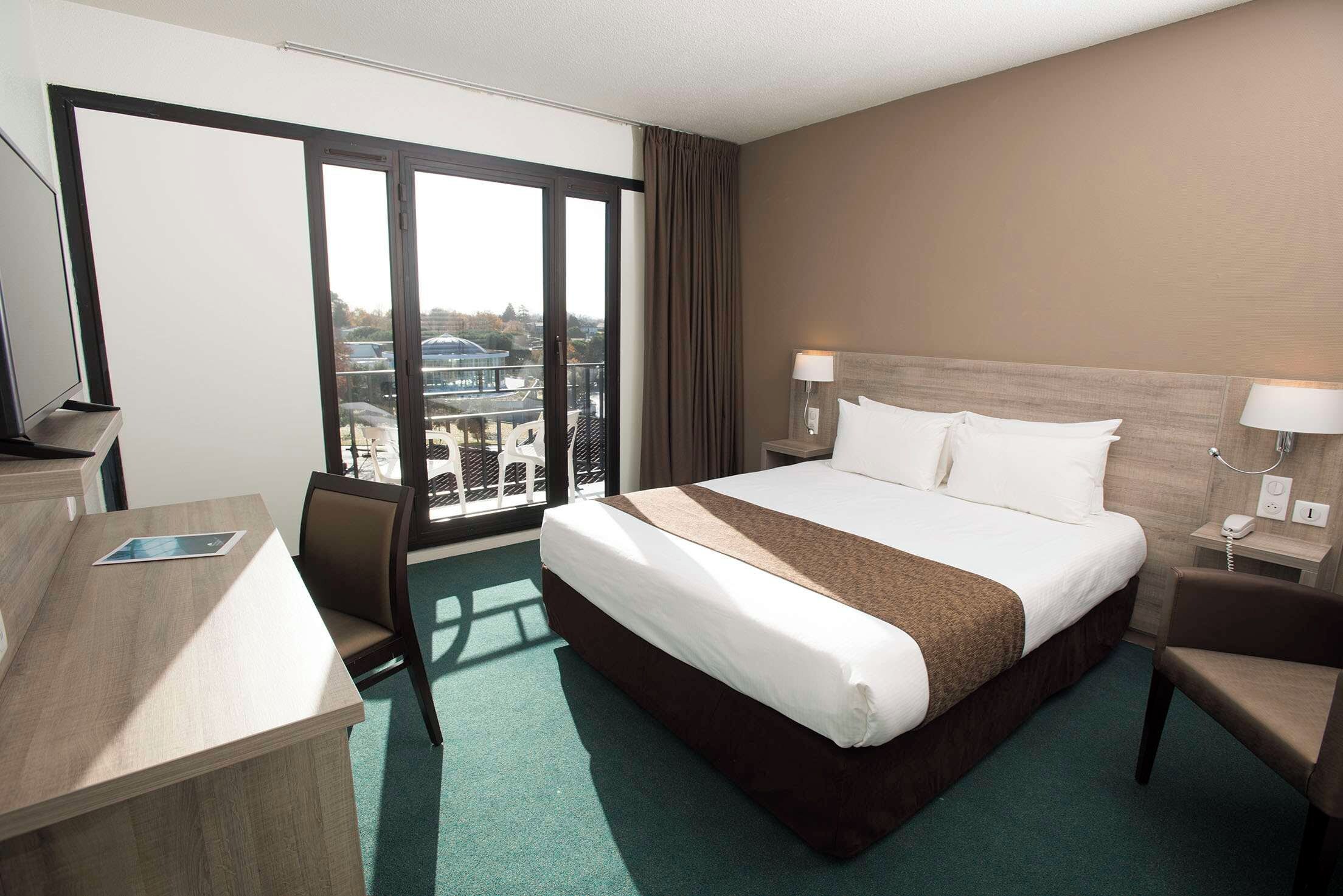 A hotel room with a large bed, a desk, and a chair. There is a view of buildings through the window.