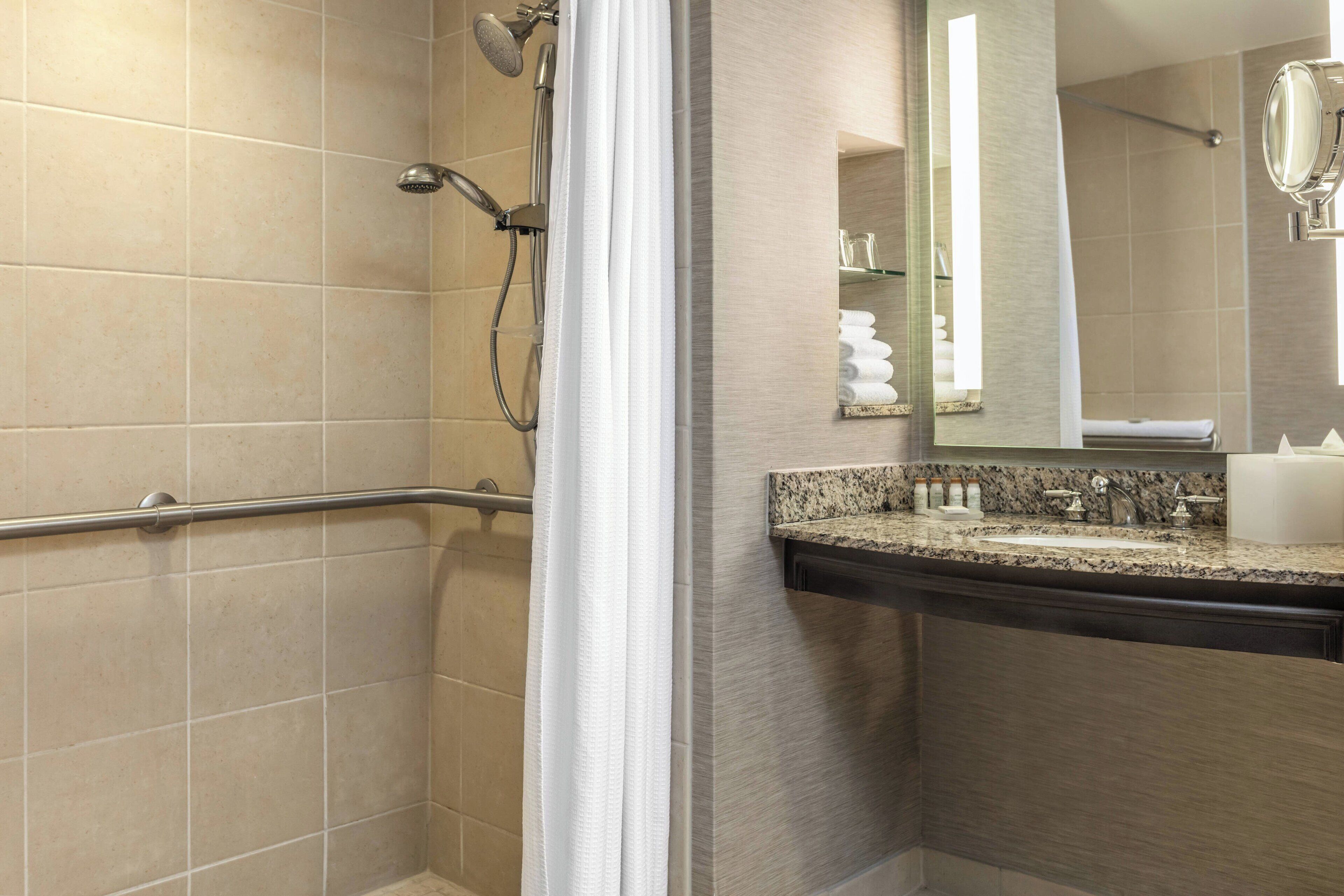 A bathroom with beige tiles, a shower with a handheld showerhead, a vanity with a granite countertop, and a mirror above the sink.
