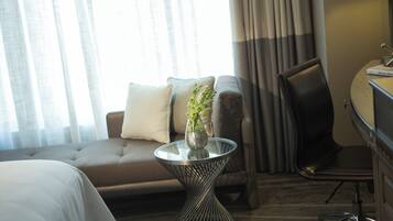 Habitación de hotel con sofá, una mesita de vidrio con un florero, una silla y un escritorio con lámpara.