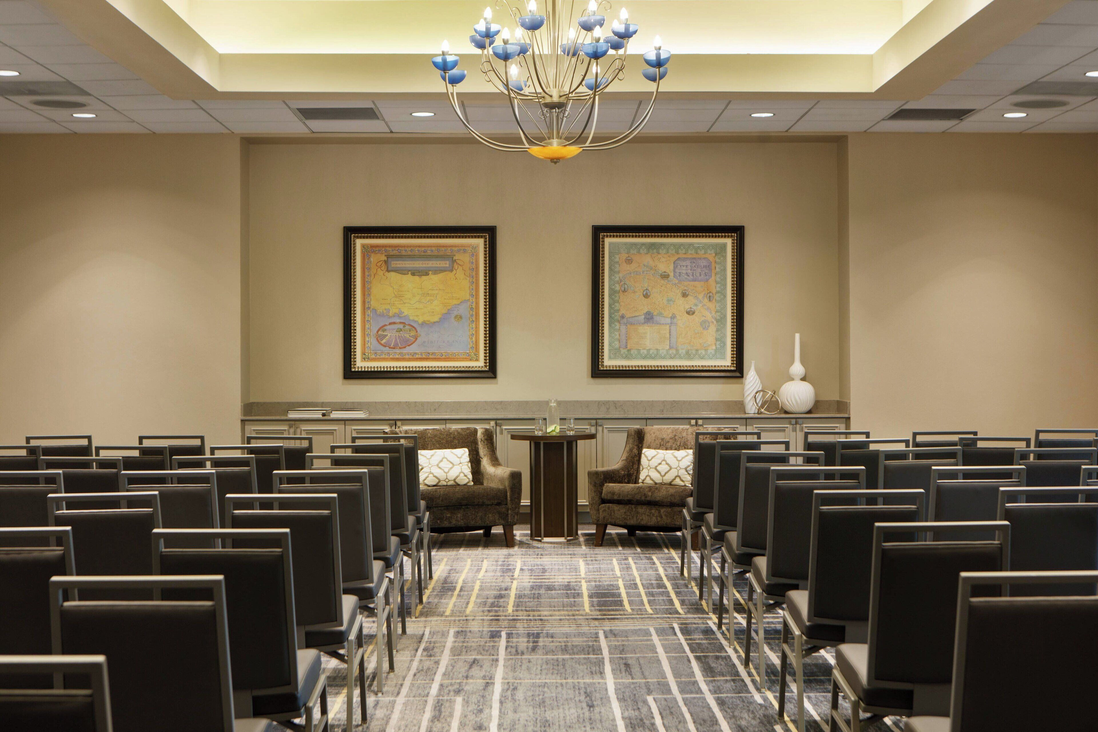 A conference room with rows of chairs, a chandelier, and two framed maps on the walls.