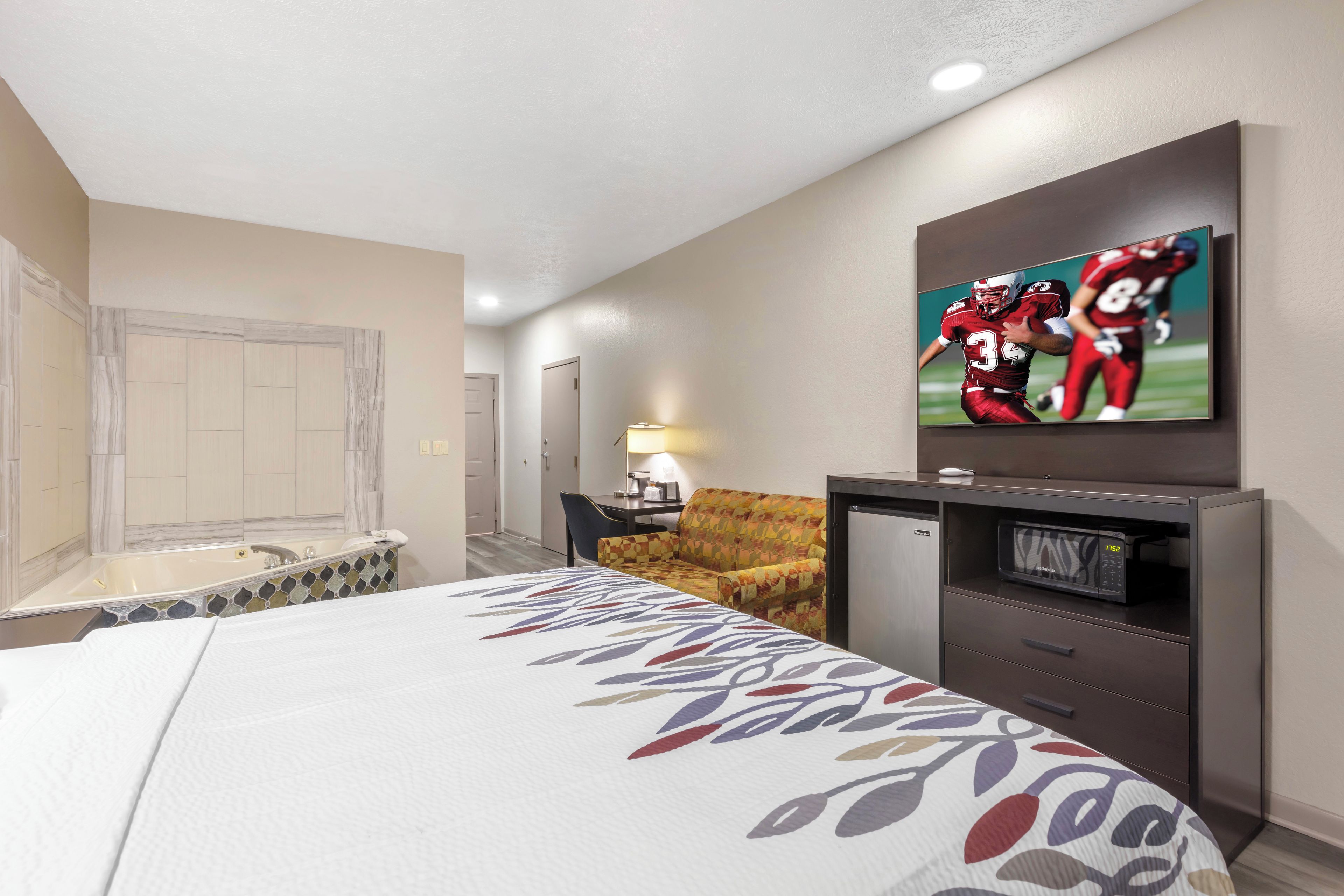 Habitación de hotel con una cama, televisión montada en la pared, escritorio con lámpara, una silla y microondas.