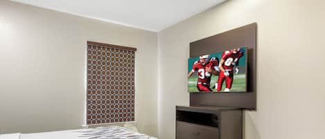 A hotel room with a bed, a TV mounted on the wall, a window with a patterned blind, and a wooden floor.