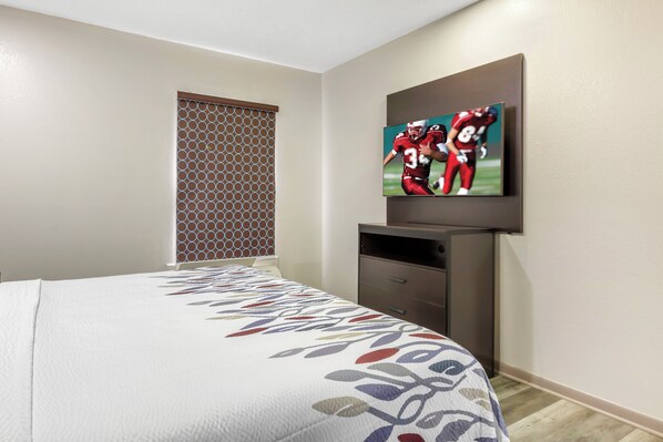 A hotel room with a bed, a TV mounted on the wall, a window with a patterned blind, and a wooden floor.