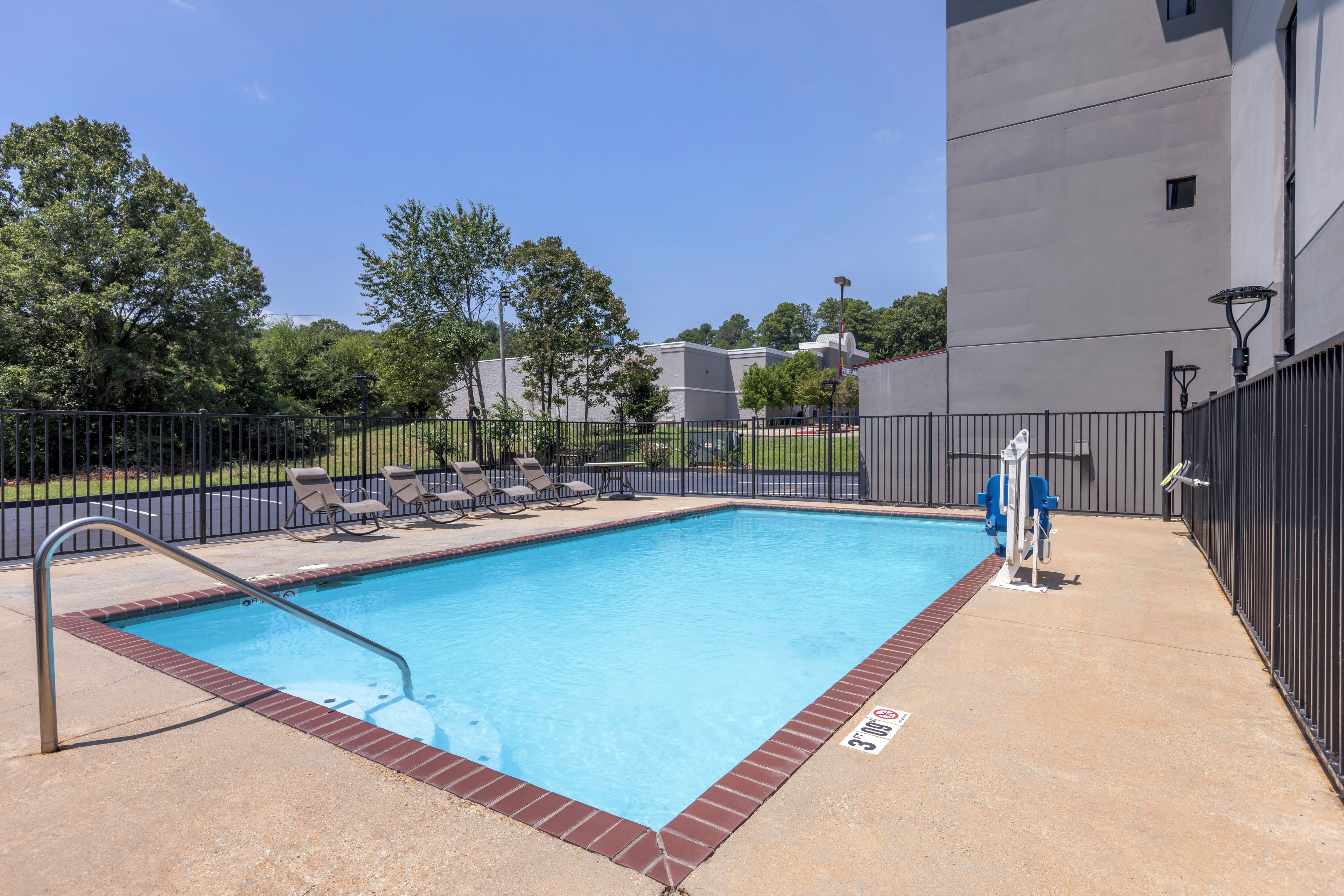 A rectangular swimming pool with a metal ladder, surrounded by a brick border and a black fence.