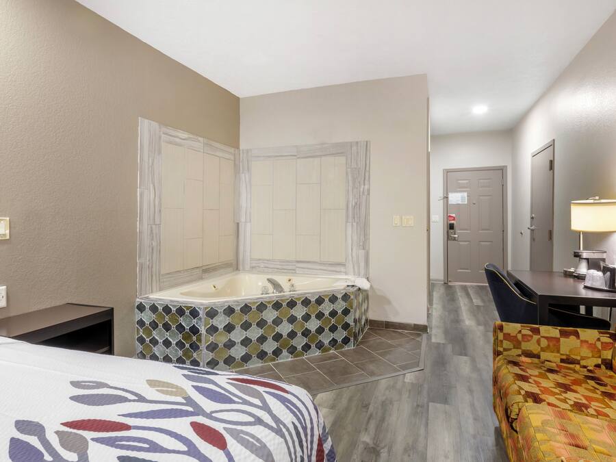 A hotel room with a large bathtub, a bed with a patterned bedspread, a chair, and a desk with a lamp.