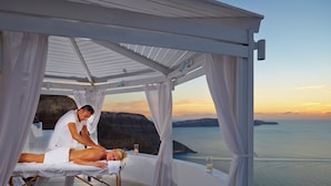 A spa treatment area with a person receiving a massage, a candle, and a glass of drink on a balcony overlooking a coastal view. - Volcano View Hotel Santorini (Santorini)