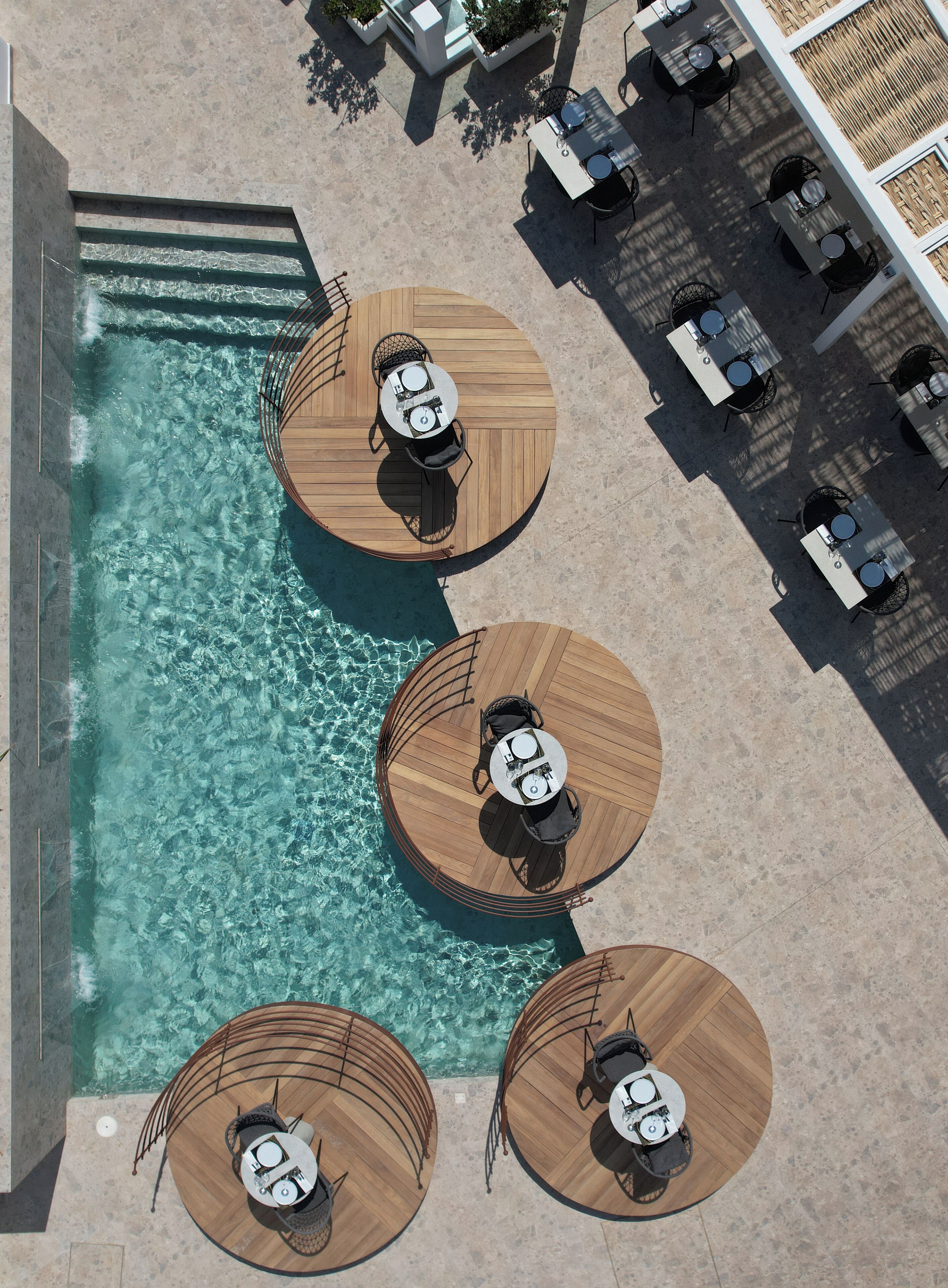 An aerial view of a poolside area with three wooden decked tables and chairs, surrounded by a tiled deck and a clear pool.
