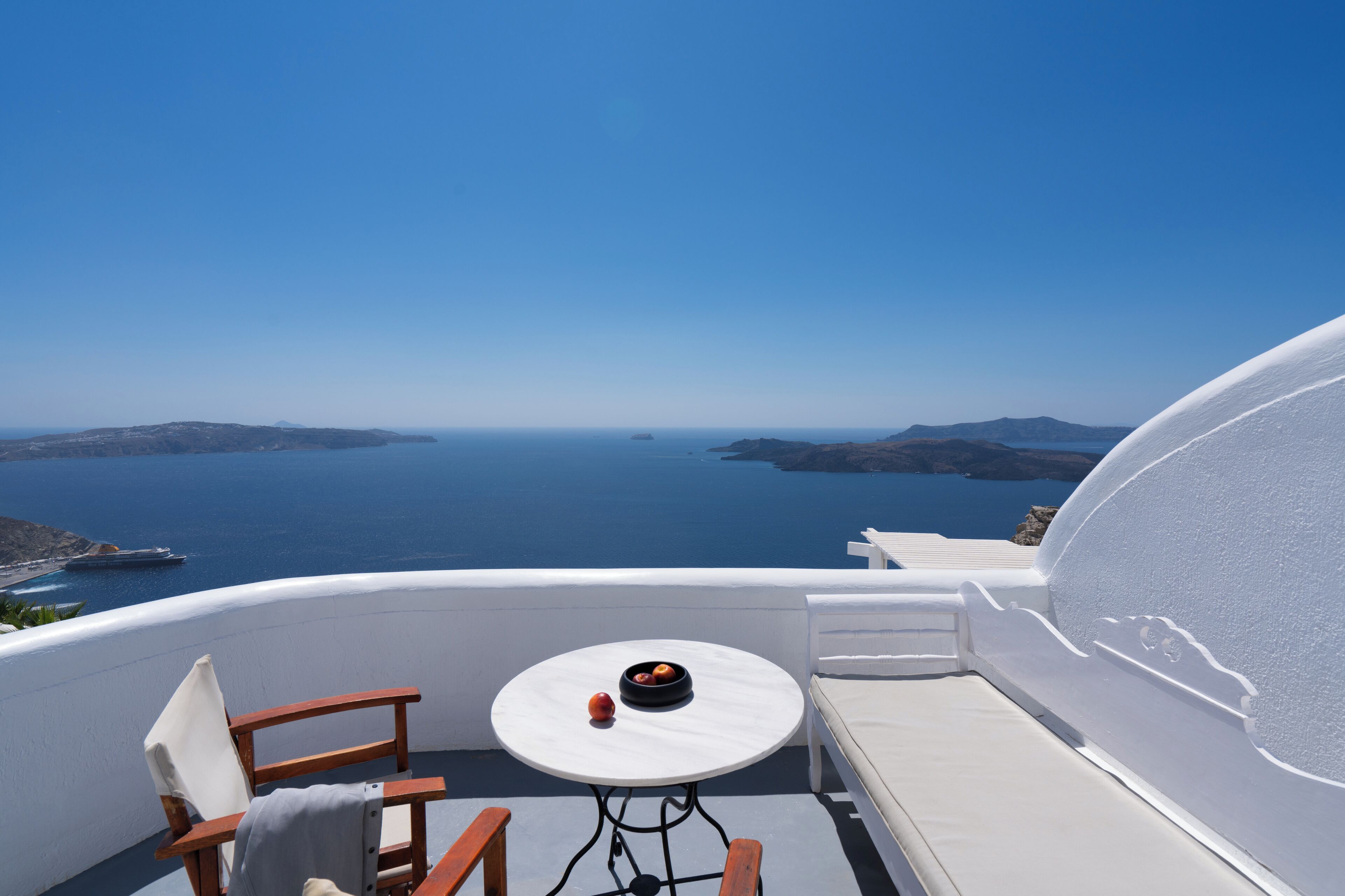 A balcony with a white table and chairs, overlooking a scenic view of the sea and distant islands.