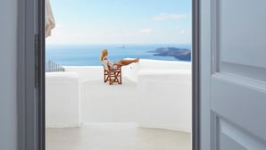 A balcony with a white interior, a wooden chair, and a view of the sea and distant land. - Volcano View Hotel Santorini (Santorini)
