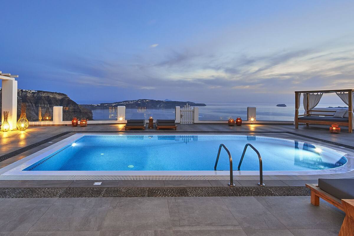 A rooftop pool with a view of the sea and mountains, surrounded by lounge areas and a wooden canopy.