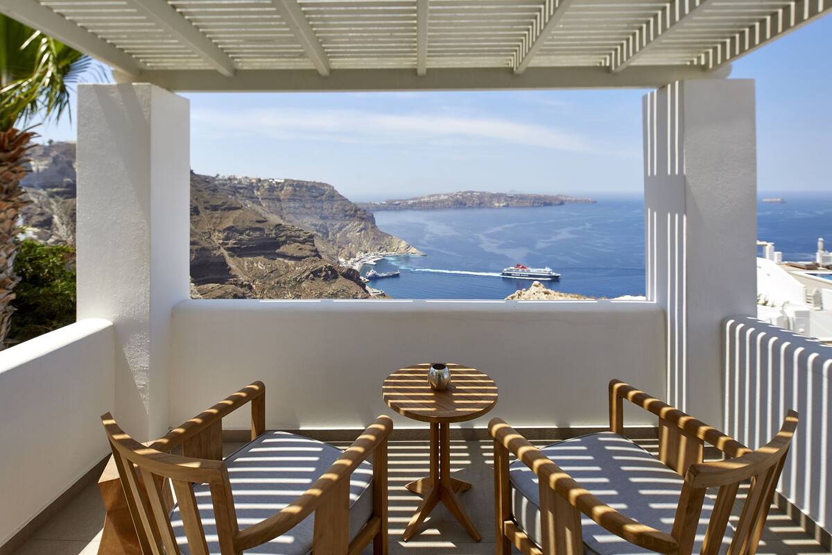 A balcony with a table and chairs, overlooking a coastal landscape with cliffs and a ship in the distance.
