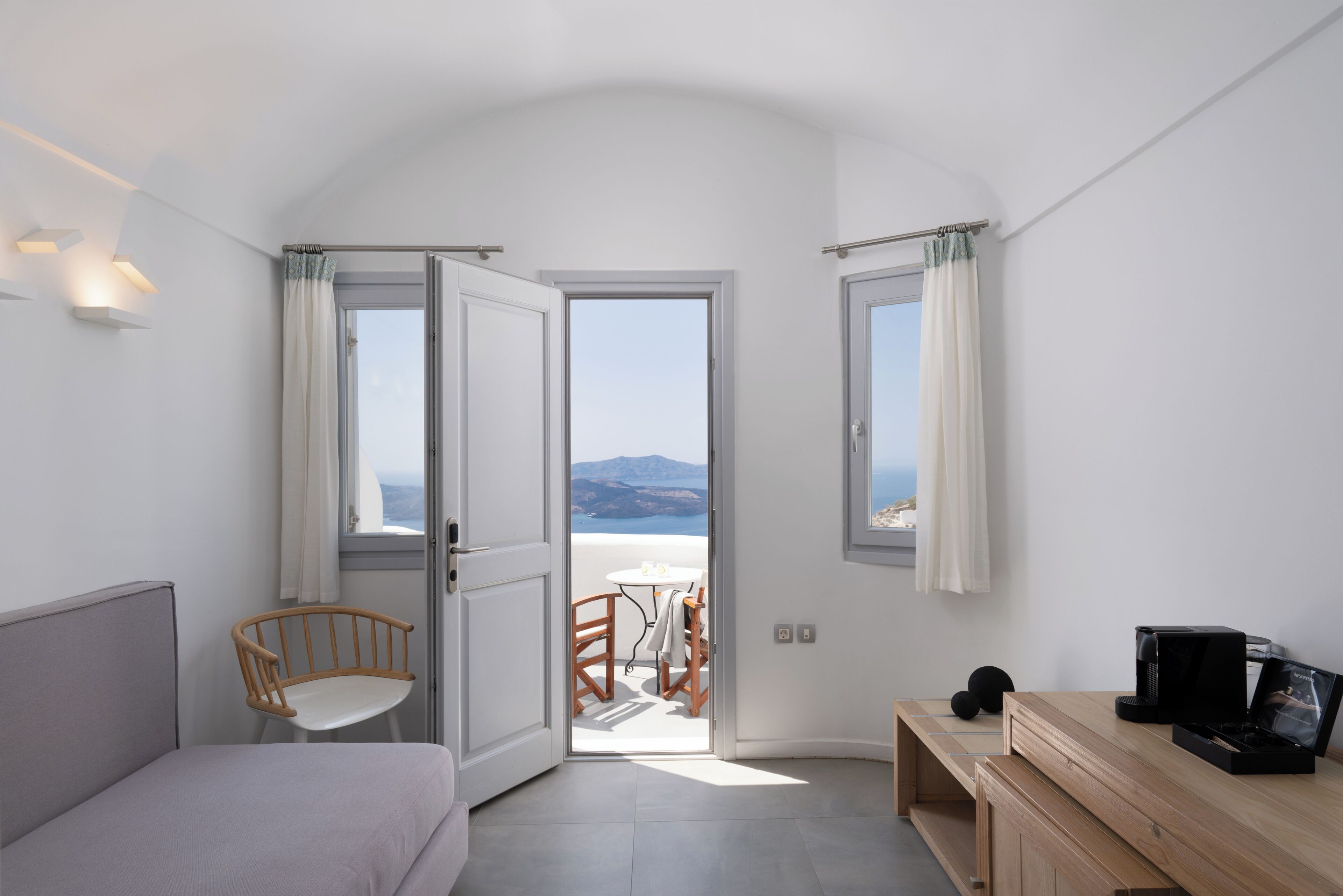 Un dormitorio con una cama, una silla de madera, un escritorio y vista al mar a través de una puerta abierta.