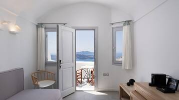 Un dormitorio con una cama, una silla de madera, un escritorio y vista al mar a través de una puerta abierta.