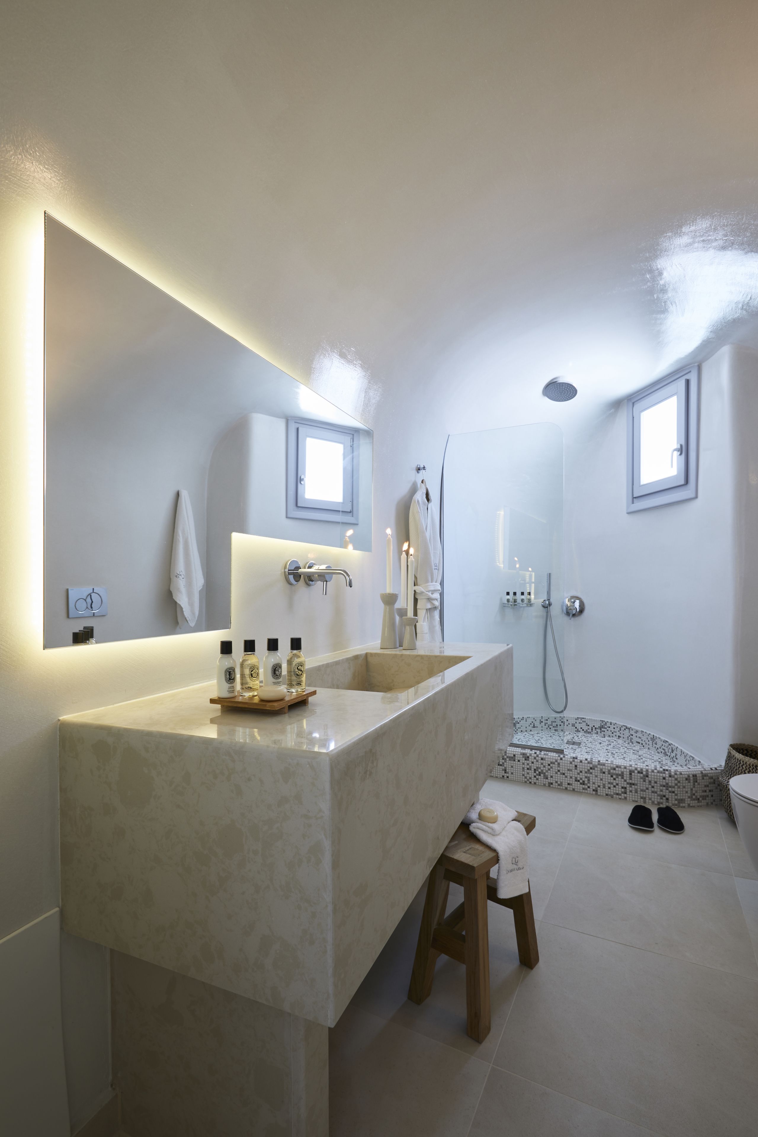 A modern bathroom with a large mirror, two sinks, a shower, and a small stool.