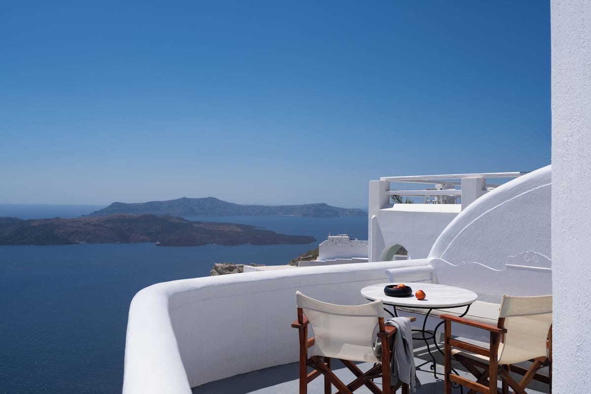 A white-walled balcony with a table and chairs overlooking a clear blue sea and distant islands.