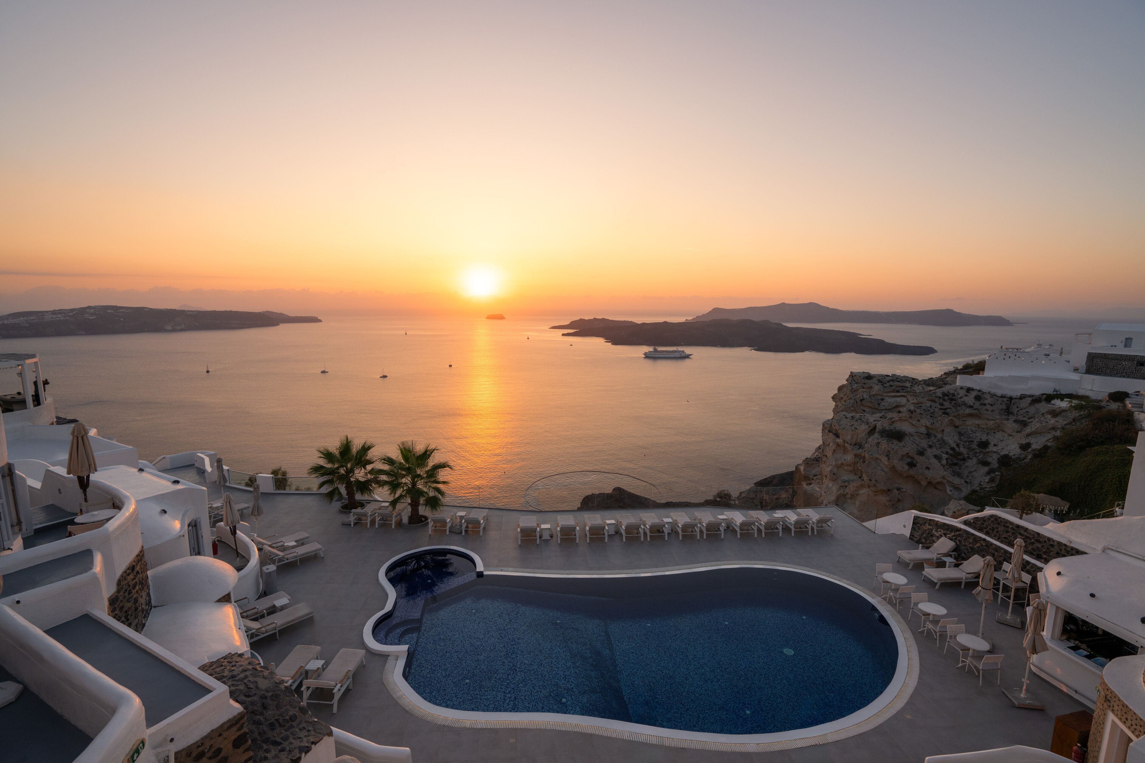 A resort with a pool, lounge chairs, and a view of the sea at sunset.