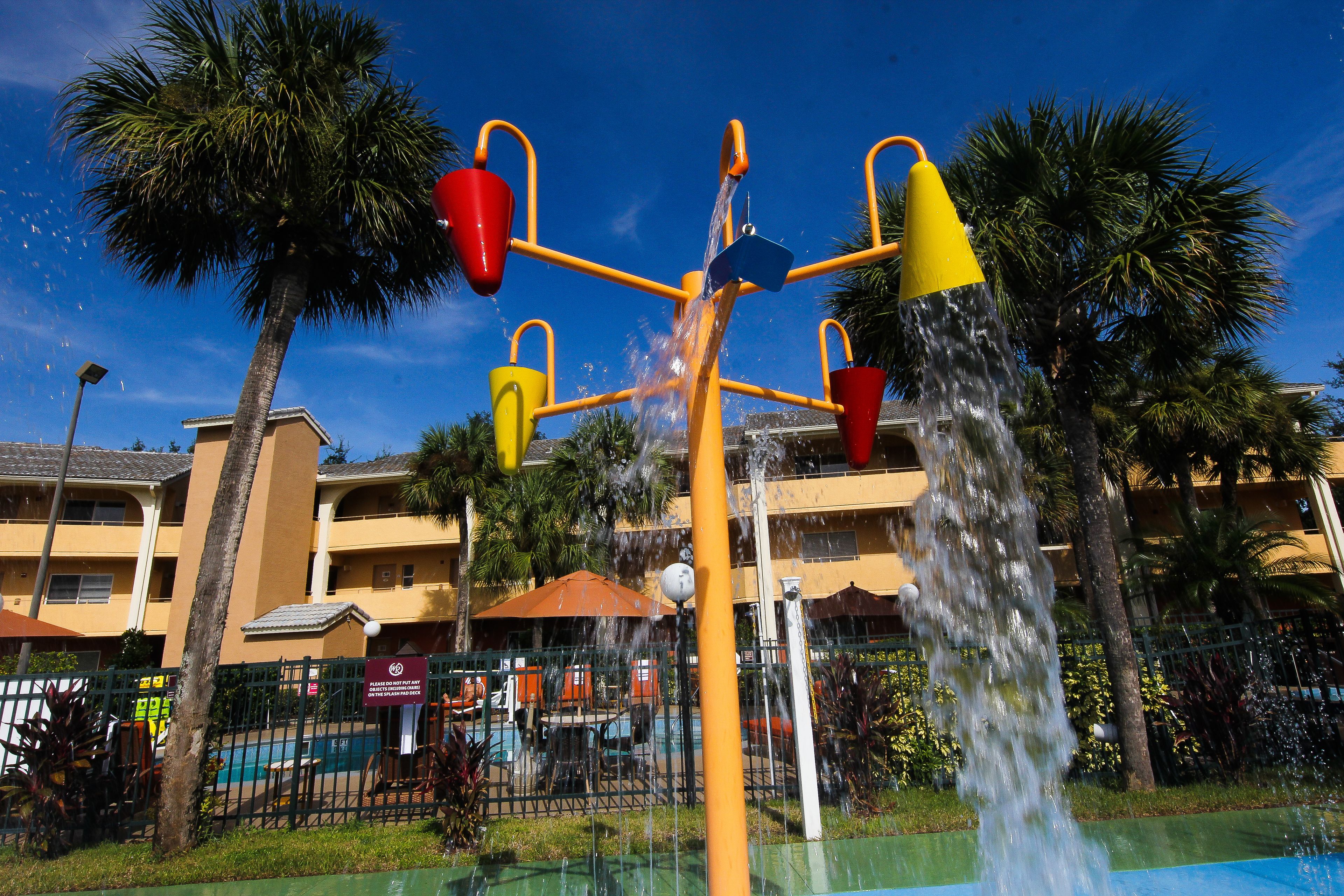 A water play area with multiple water jets, palm trees, and a pool in the background.