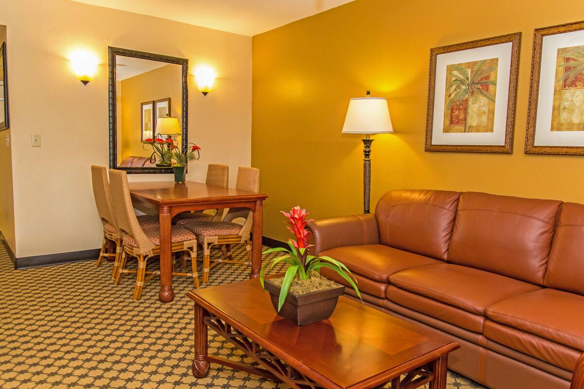 A hotel room with a dining table, chairs, a sofa, a lamp, and two framed pictures on the wall.