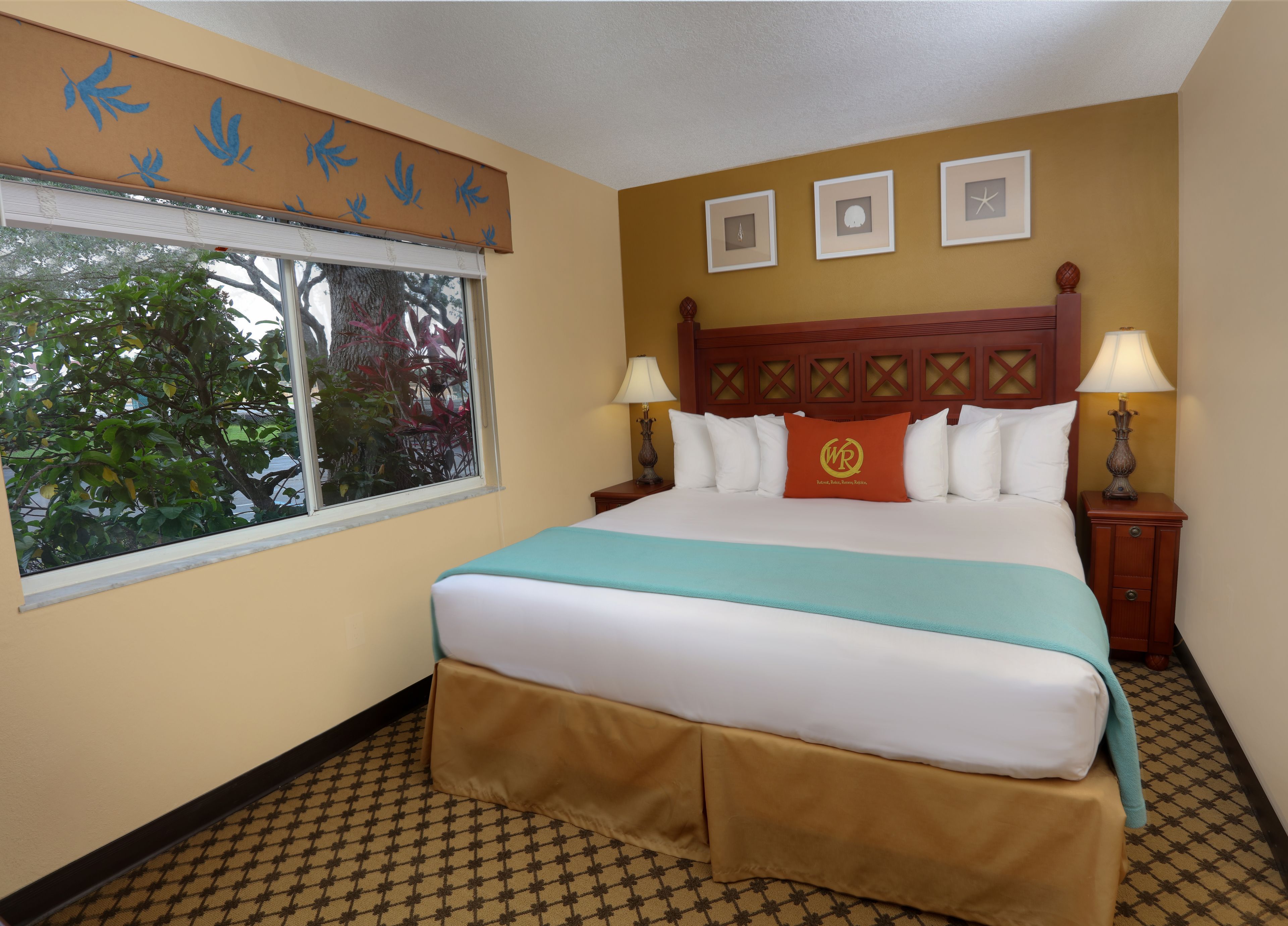 A hotel room with a large bed, two bedside tables with lamps, a window with a view of trees, and three framed pictures on the wall.